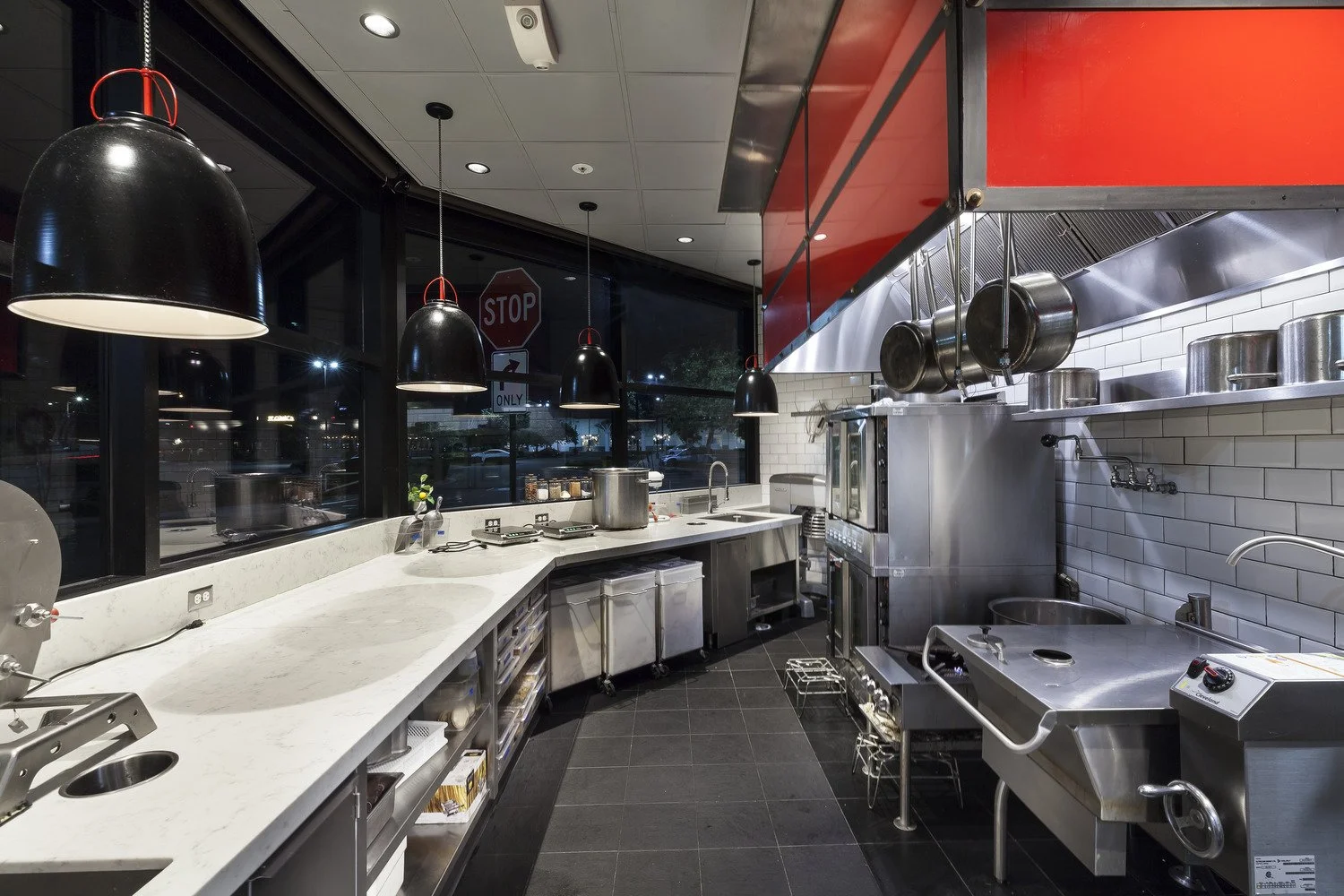 Empty professional commercial kitchen with black pendant lights, white marble countertops, stainless steel appliances, pots hanging above stove, white subway tile backsplash, and large windows showing a night scene outside.