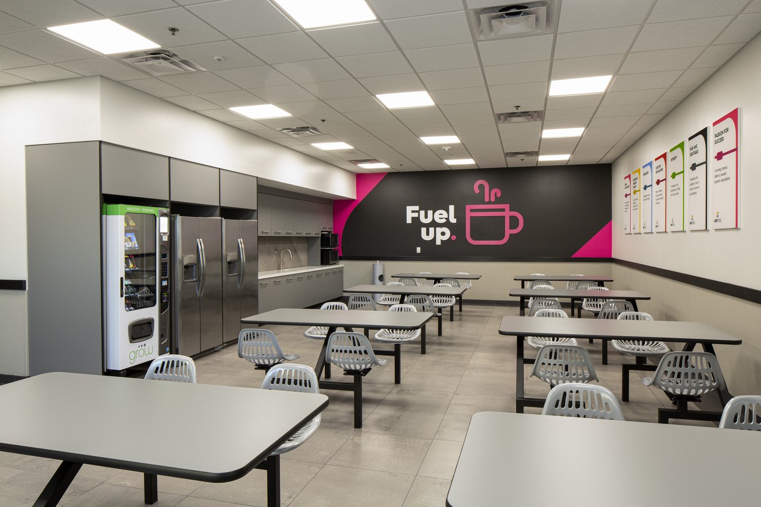 An empty cafeteria with white tables and gray chairs, a beverage vending machine, and a wall with a pink and black mural that reads "Fuel up" with a pink coffee cup icon.