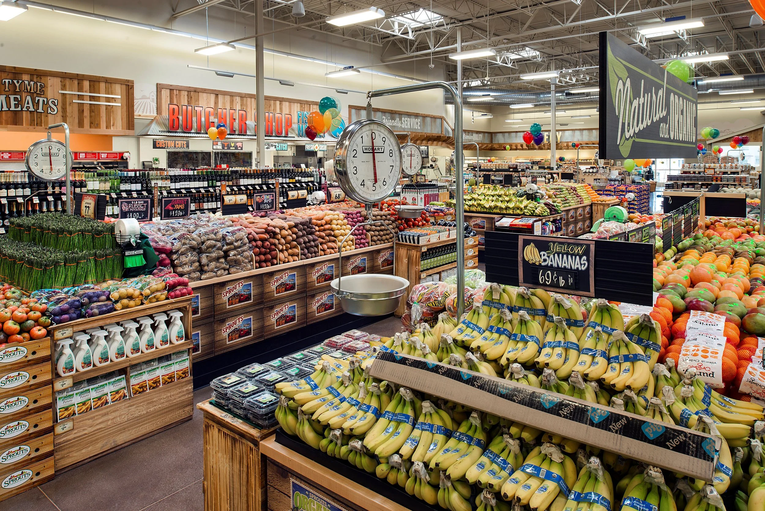 Inside a grocery store with fresh produce including bananas, tomatoes, sweet potatoes, and onions. There are clocks hanging above the produce section, and a large sign that reads "Natural and Organic". Other store signs include "Butcher Shop" and "Me