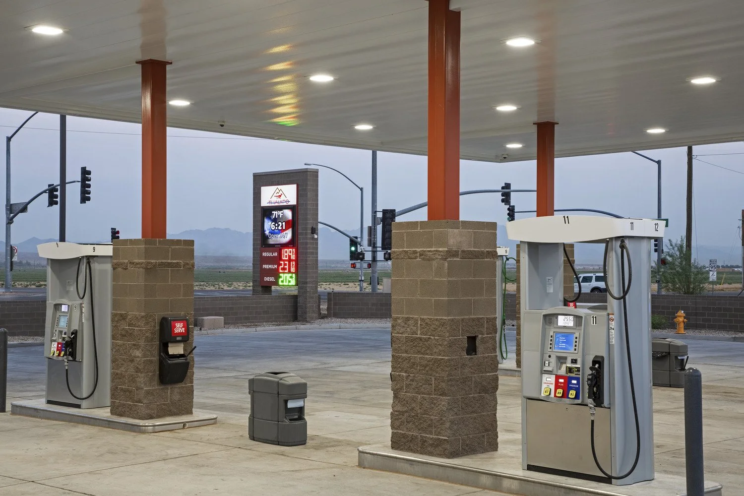Gas station with fuel pumps, a digital price sign, and a background of traffic lights and mountains.