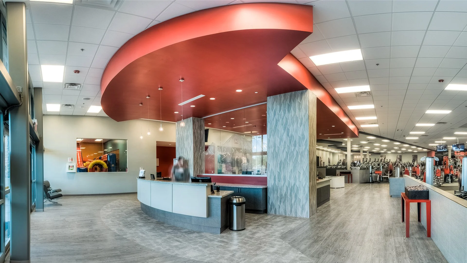 Modern gym reception area with a curved white counter, red ceiling, and workout equipment visible in the background.