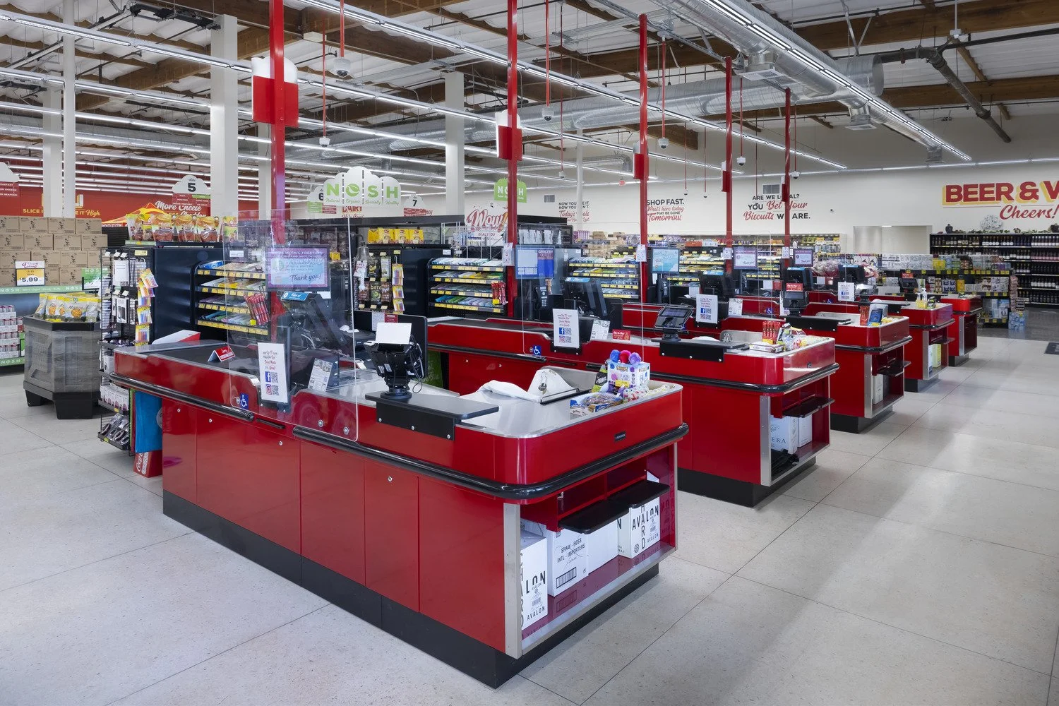 Grocery store checkout counters with red and black design, cash registers, payment stations, and plastic shields, in a spacious store with shelves of products and signage in the background.