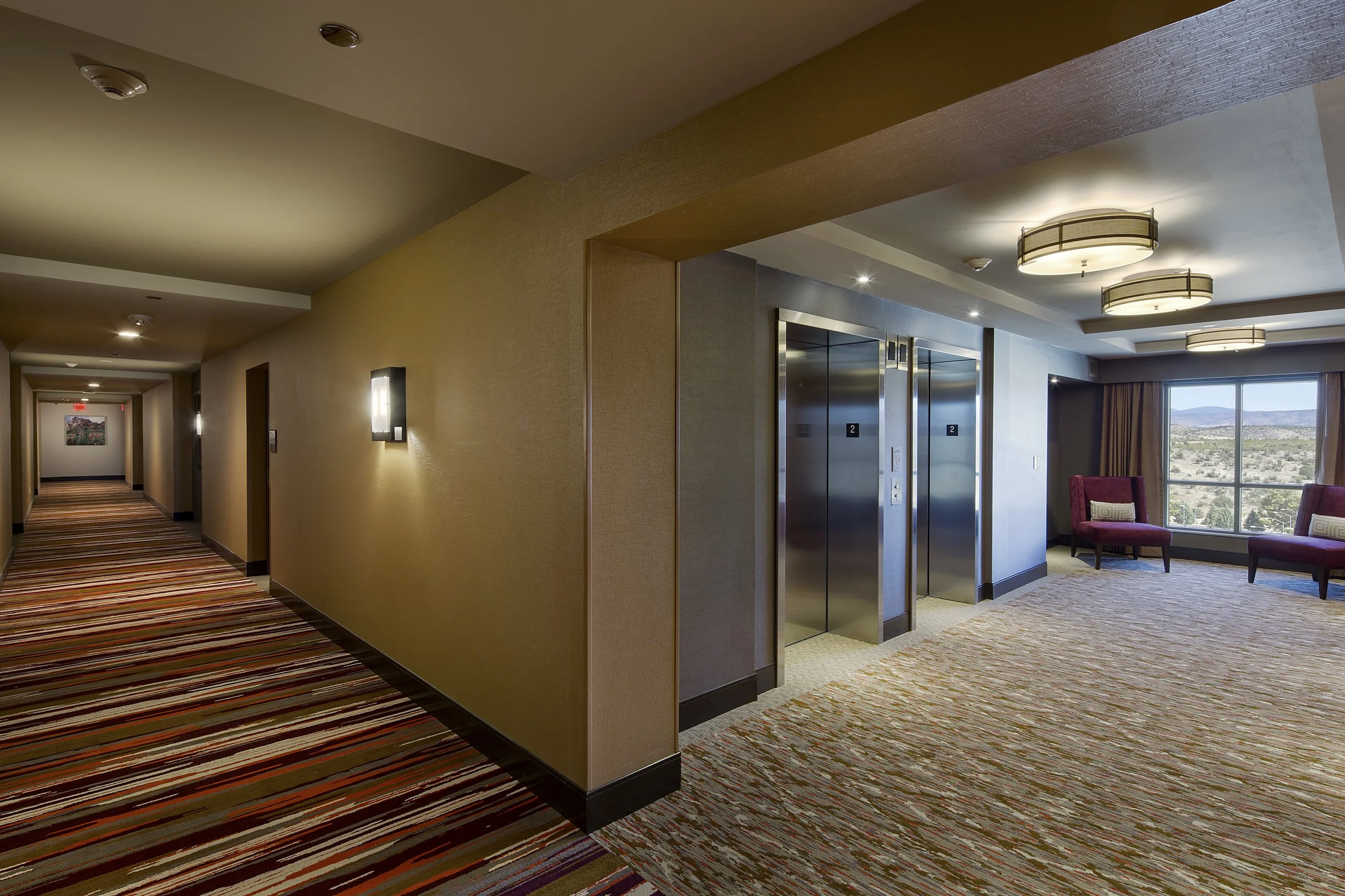 Hotel corridor with an elevator lobby area, two purple chairs near a large window overlooking a landscape, and colorful striped carpet.