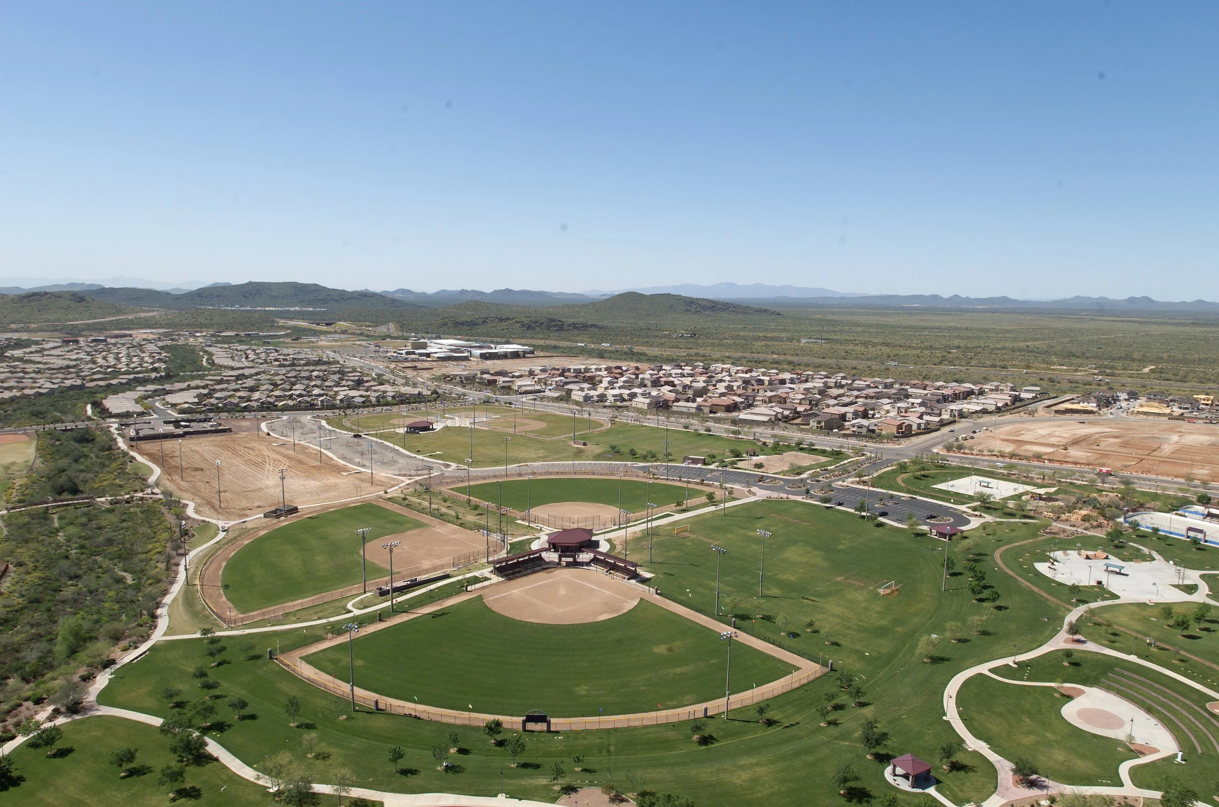 Aerial view of a community park with baseball fields, walking paths, and a residential neighborhood in the background under a clear blue sky.
