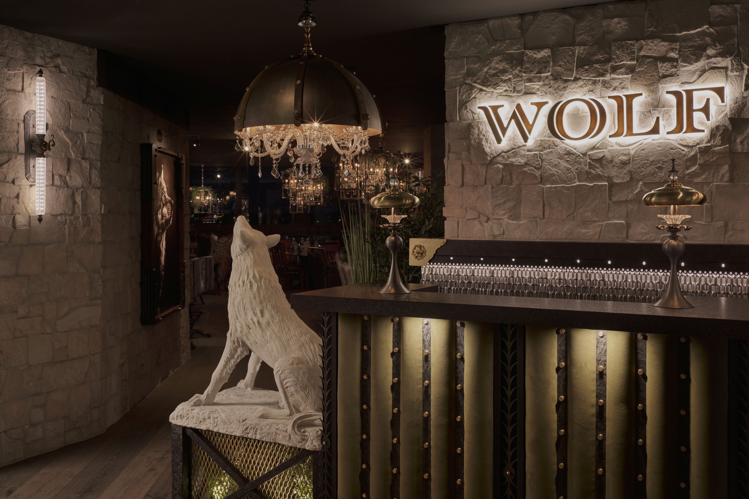 Interior of a restaurant with a stone wall, illuminated sign reading 'WOLF', a wolf sculpture on a stand, chandeliers, and elegant lighting fixtures.