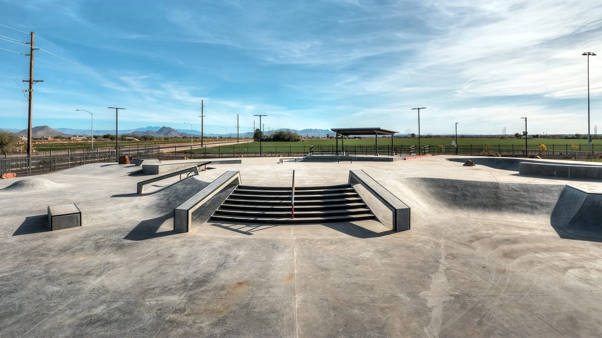 An empty outdoor skatepark with various ramps, stairs, and rails under a blue sky with clouds, in a rural area with mountains in the distance.