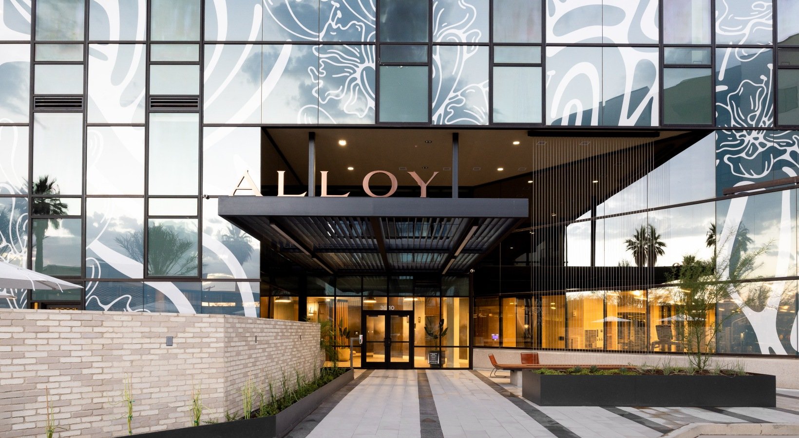 Modern building with large reflective glass windows, decorative white patterns, and a sign that reads 'ALLJOY' above the entrance, with planters and benches outside.