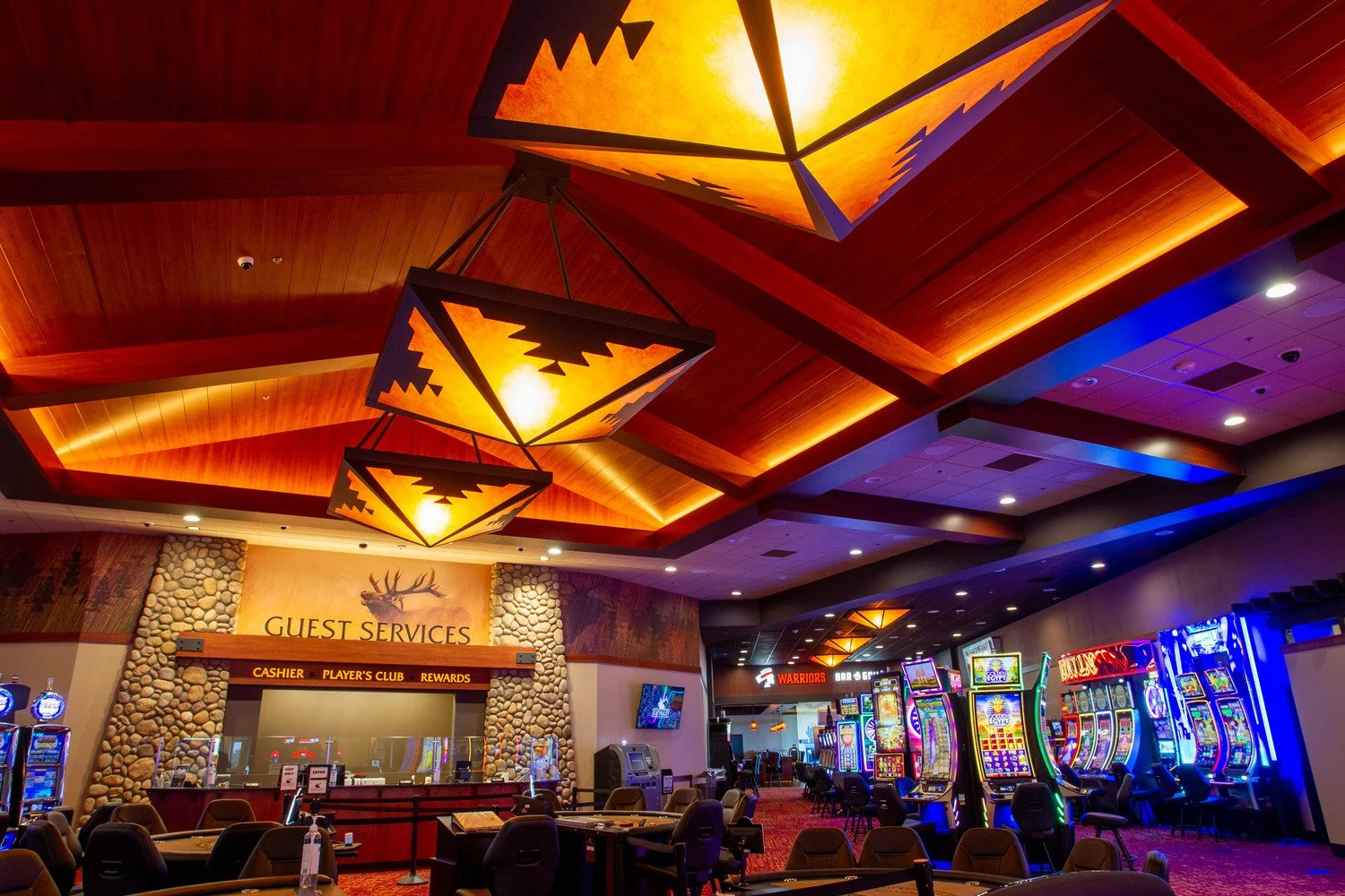 Inside a casino with slot machines, a guest services desk, and a wooden ceiling with geometric light fixtures