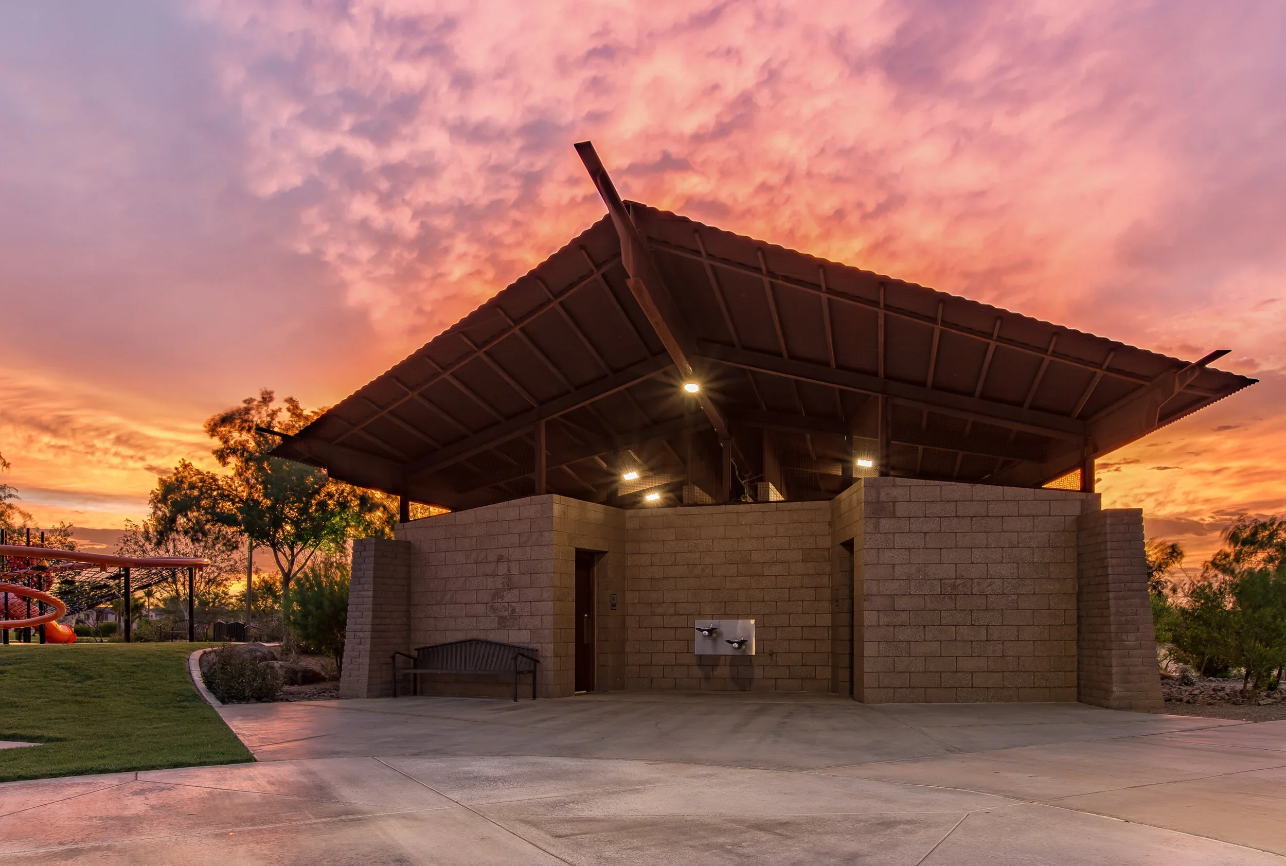 Public restroom building with a sloped roof at sunset, surrounded by trees, with a bench and a playground slide visible nearby.