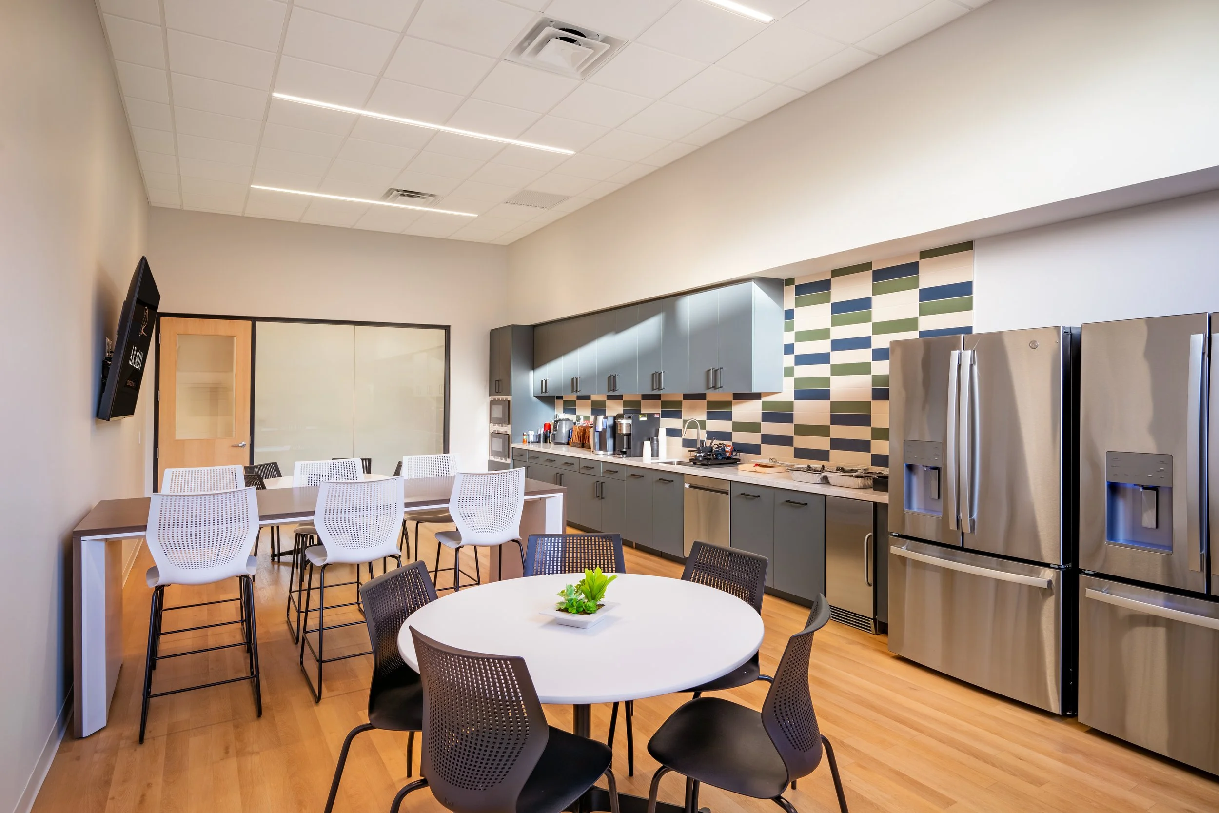 Modern break room or kitchenette with stainless steel appliances, a round table with black chairs, a counter with cabinets, and colorful tile backsplash.