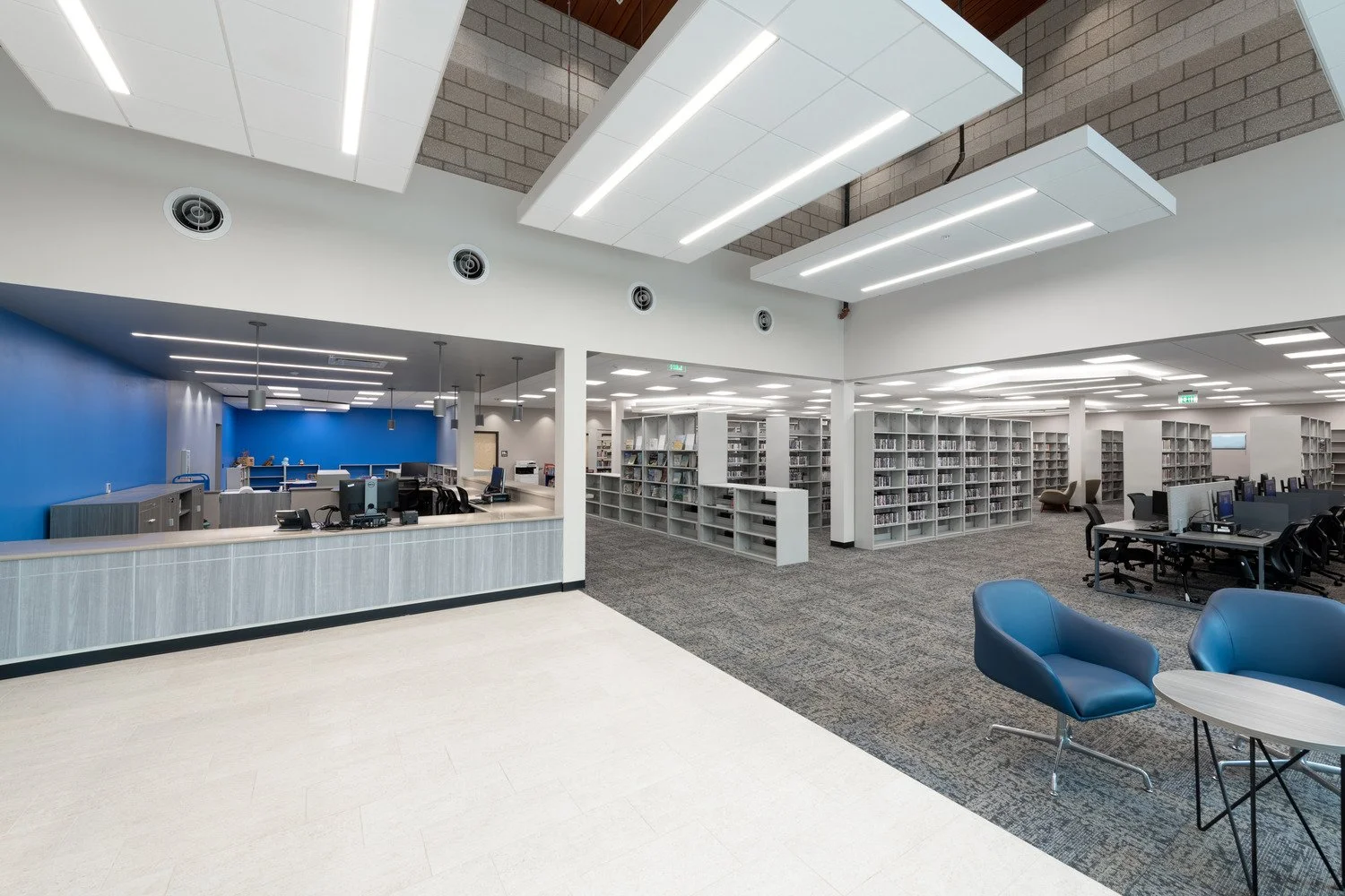 Interior of a modern library with a reception desk, rows of bookshelves, study areas with computers and chairs, and bright lighting.
