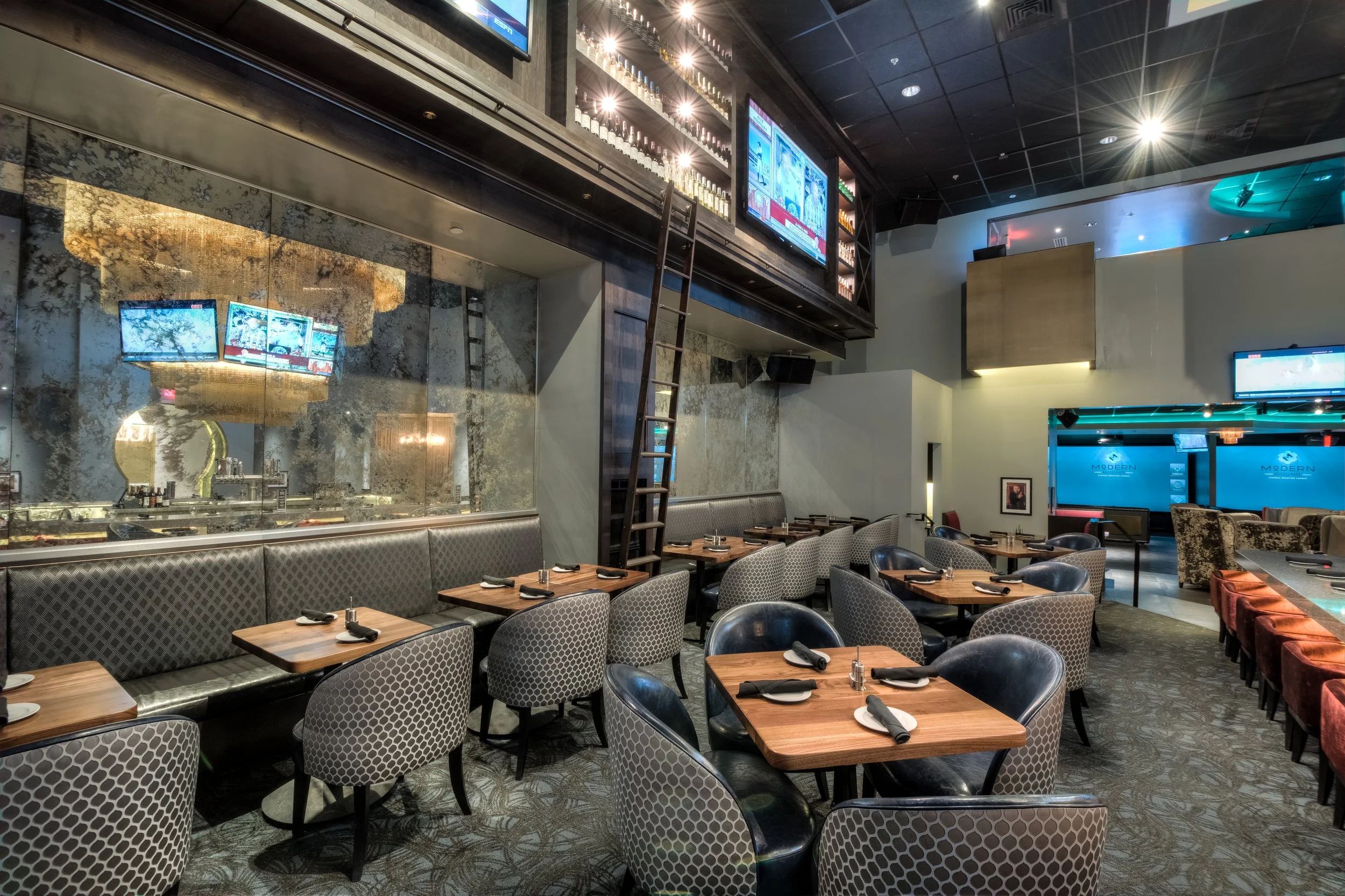 Upscale restaurant interior with dining tables set with black cloth napkins and white plates. Wall-mounted TVs and modern decor throughout.