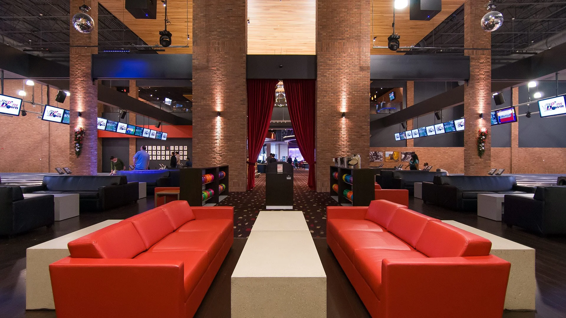 Interior view of a bowling alley with red and black couches, bowling lanes, and multiple screens displaying scores and graphics. Brick walls and red curtains are also visible.