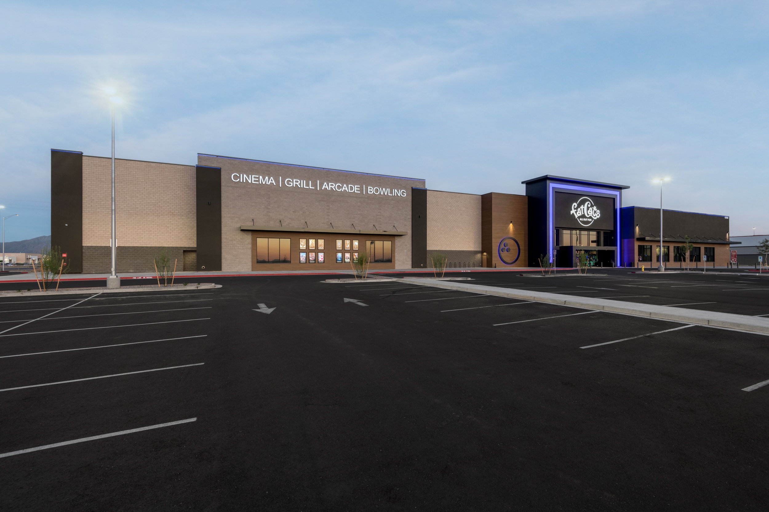 A large commercial building with a parking lot in front. The building has signs indicating amenities like cinema, grill, arcade, and bowling. The entrance is illuminated with purple lighting, and there are a few street lamps and small plants in the p