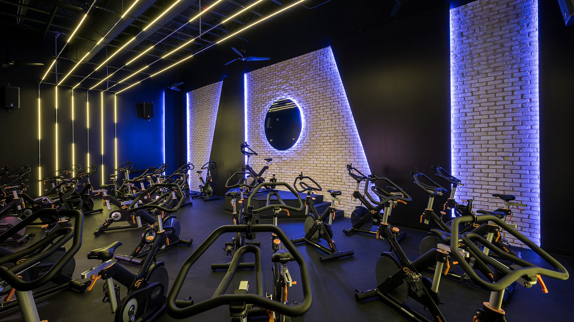 Indoor spin class room with black stationary bikes, modern purple and yellow LED lighting, exposed brick wall with a circular mirror, and ceiling fans.