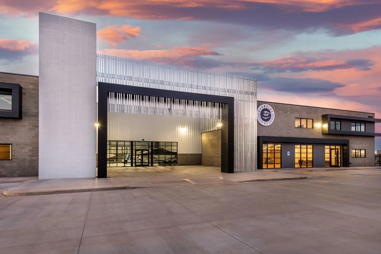 View of a modern warehouse or industrial building at sunset, with a large open entrance, garage doors, and signage reading 'GERMANFIRE' on the exterior, and a spacious parking area in front.