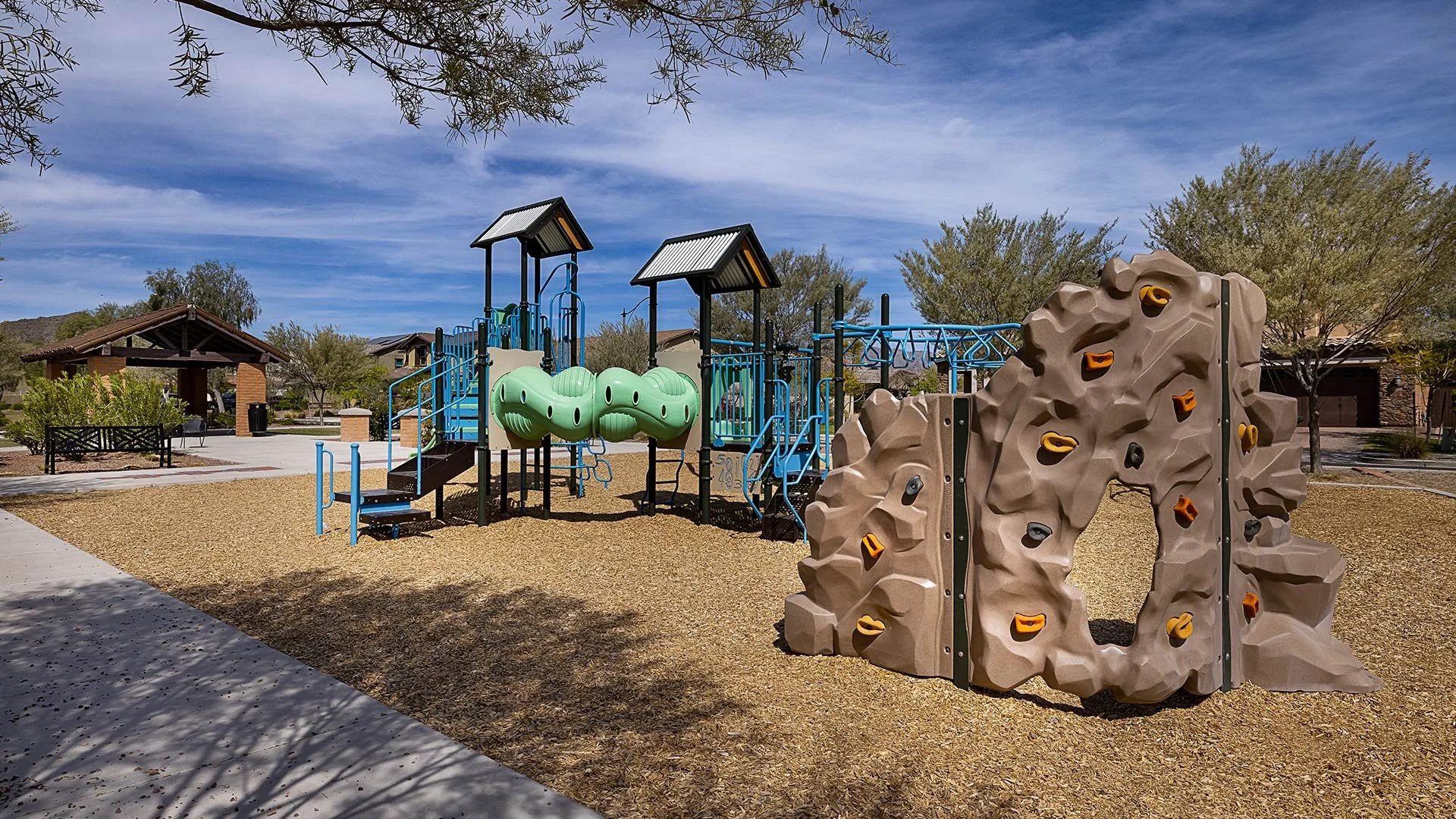 Empty playground with climbing structures, slide tunnels, and a rock climbing wall, surrounded by trees and a sidewalk.