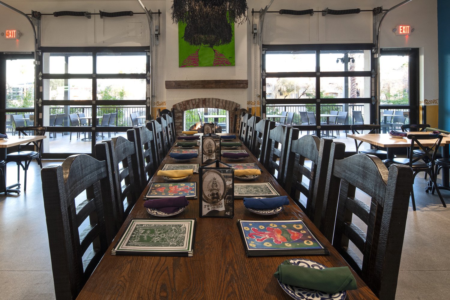 A long wooden dining table set with colorful napkins and placemats, with menus in the center, inside a restaurant with large windows, a brick fireplace, and artwork on the wall.