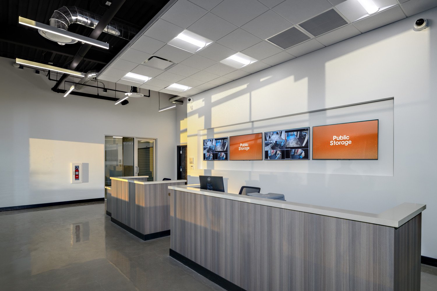 Empty reception desk area with a computer, two chairs, and multiple security camera monitors on the wall displaying security footage. The room has bright overhead lighting, a white wall, and ventilation ducts on the ceiling. Shadows from window blind