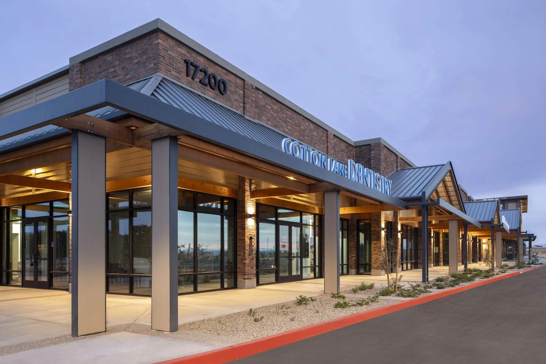 A modern commercial building housing Cotton Lane Dentistry with glass front doors, brick and metal siding, illuminated signage, and a small landscaped area outside.