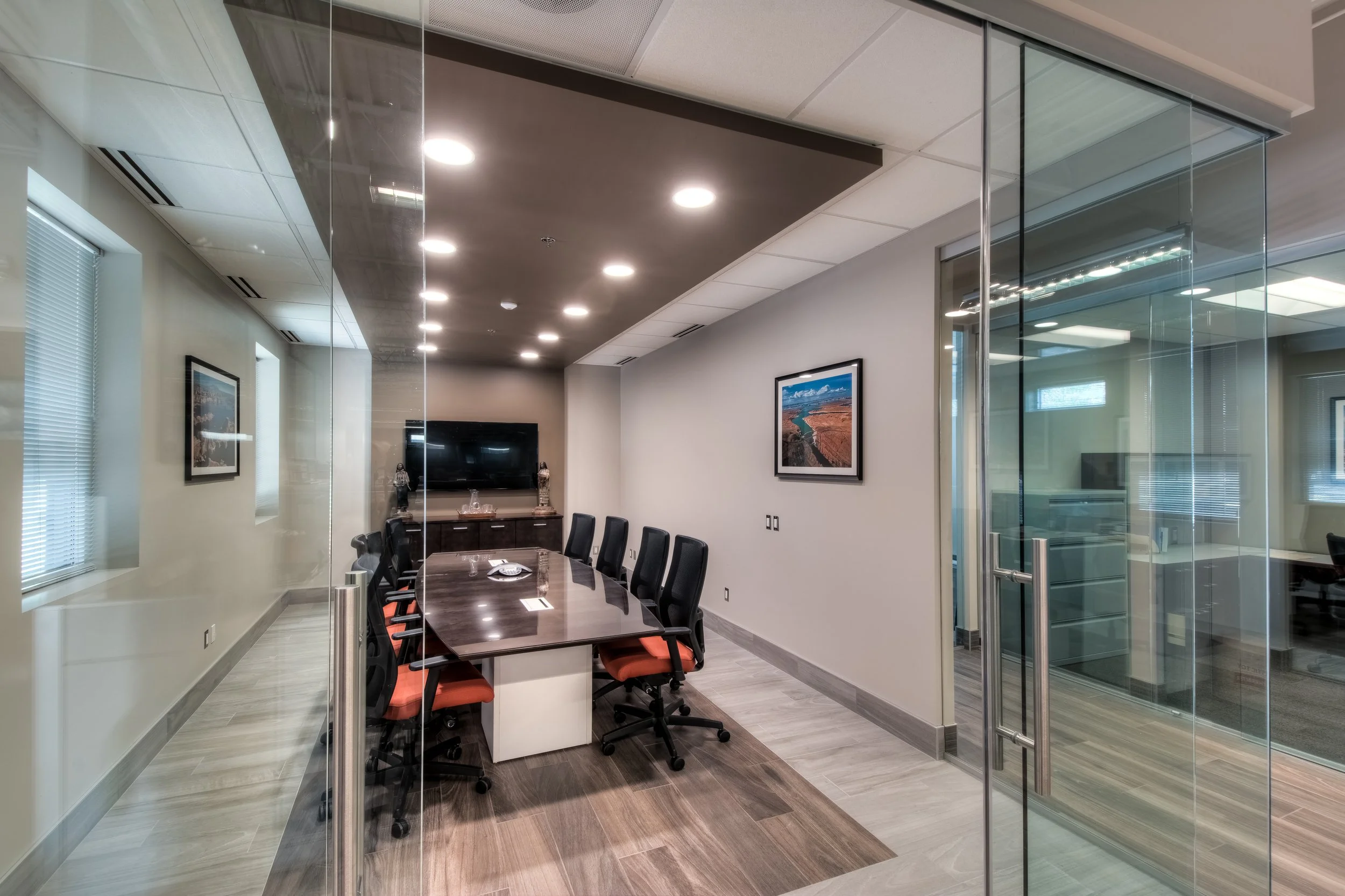 Modern conference room with a large wooden table surrounded by black chairs with orange cushions, a wall-mounted flat-screen TV, framed landscape photos on the walls, a glass door, and windows with blinds, illuminated by ceiling lights.