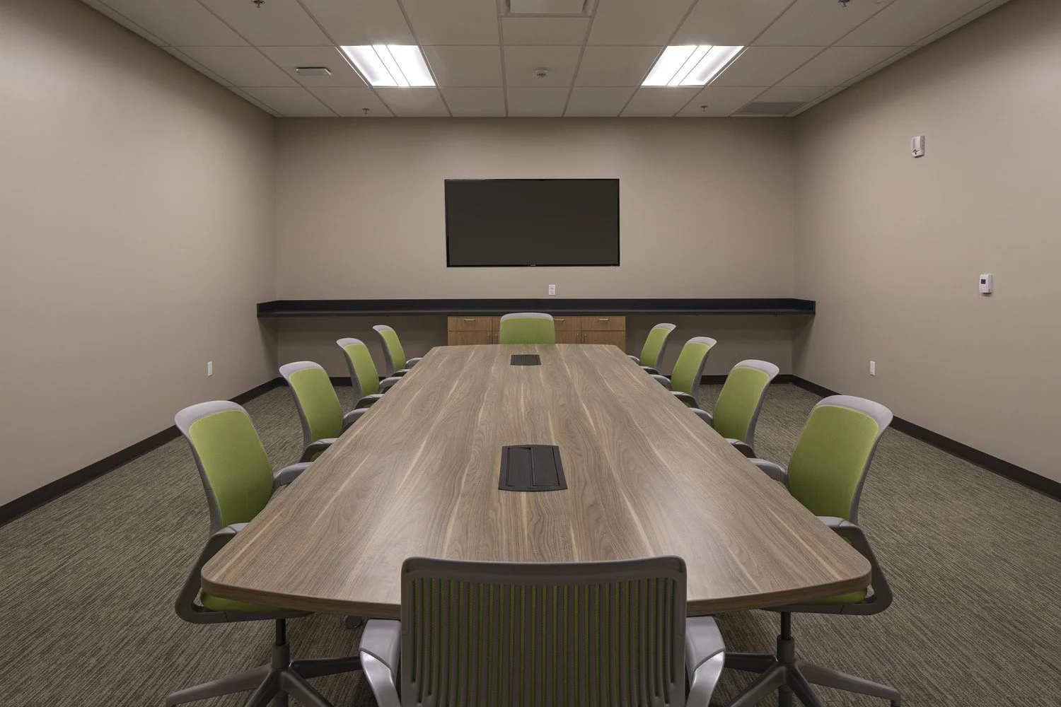 Empty conference room with a large wooden table, green chairs on both sides, a black wall-mounted screen, and beige walls.
