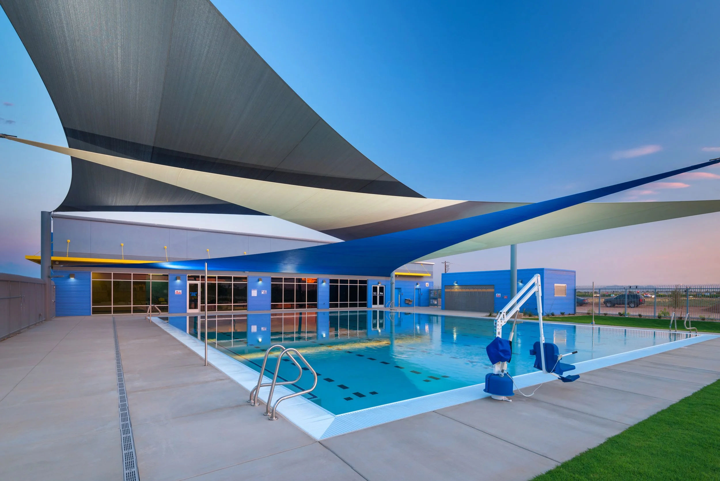 Outdoor swimming pool with a shade canopy, surrounded by a concrete deck, in front of a blue building with large windows, under a clear sky at dusk.