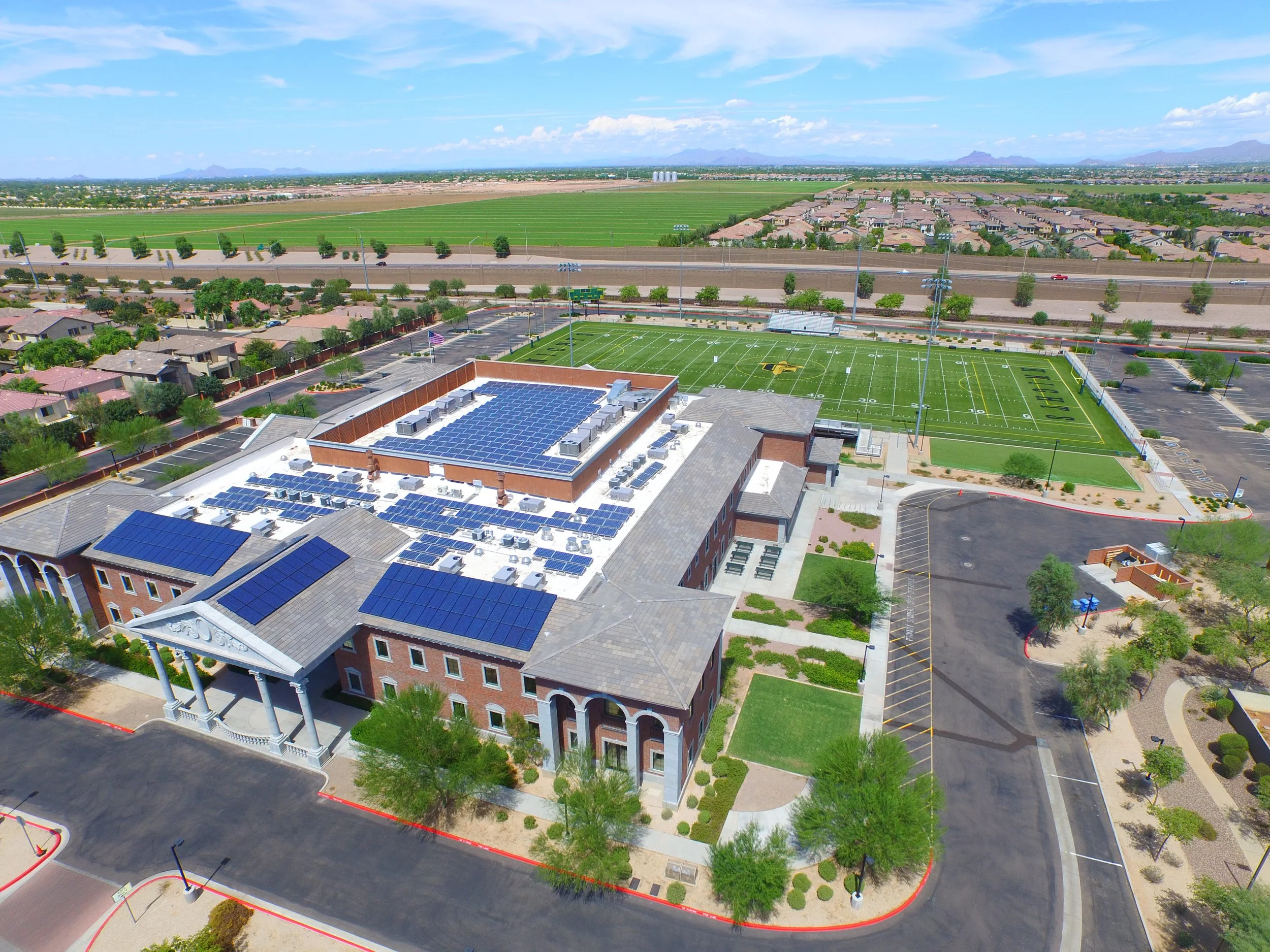 Aerial view of a building with solar panels, parking lot, a football field, and surrounding residential and rural areas under a partly cloudy sky.