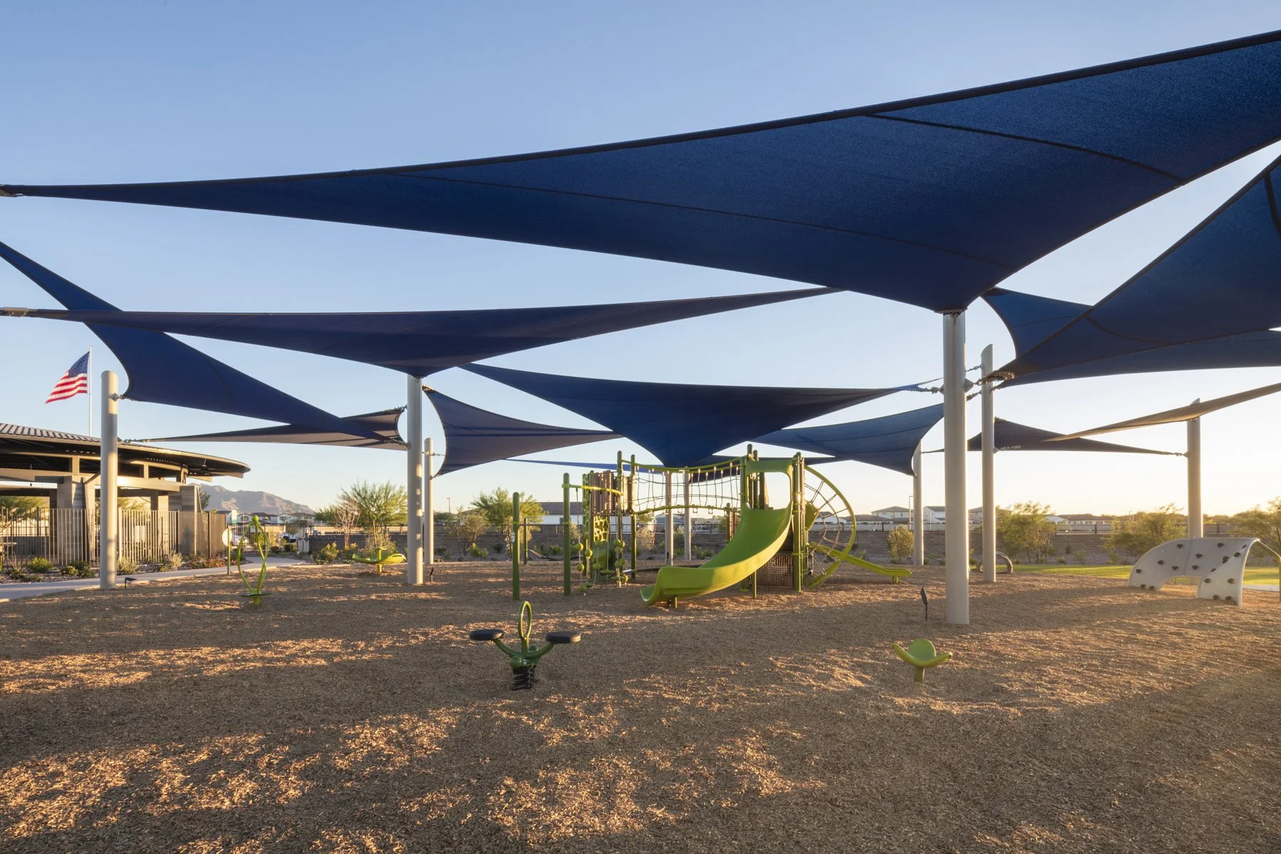 A kids' playground area with a green slide, climbing structures, and shade sails overhead, in a desert landscape at sunset.