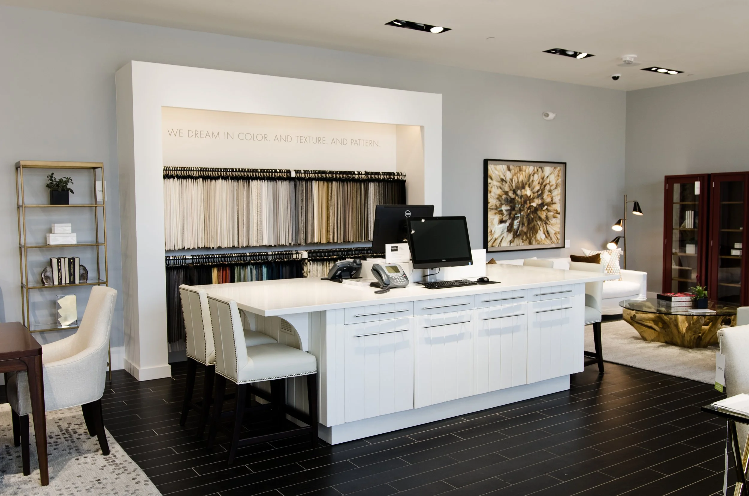 Interior view of a modern home decor store showcasing fabric swatches, a reception desk with computers and phones, a seating area with a white sofa and decorative pillows, a gold coffee table, a framed artwork, and bookshelves.