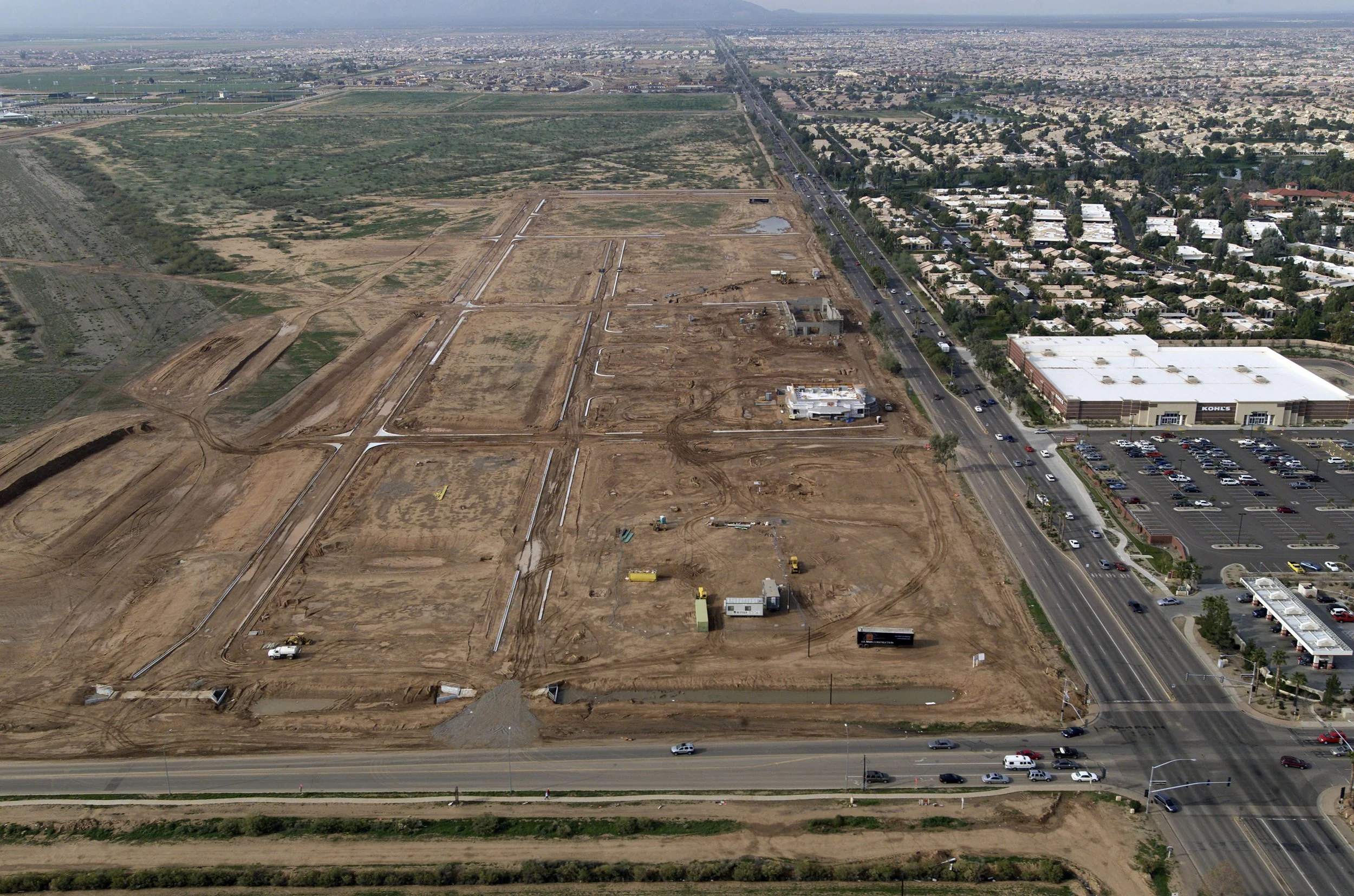 An aerial view of a construction site with dirt and gravel, roads, machinery, and some structures in progress. To the right, there is a commercial area with a parking lot, retail stores, and nearby residential neighborhoods.