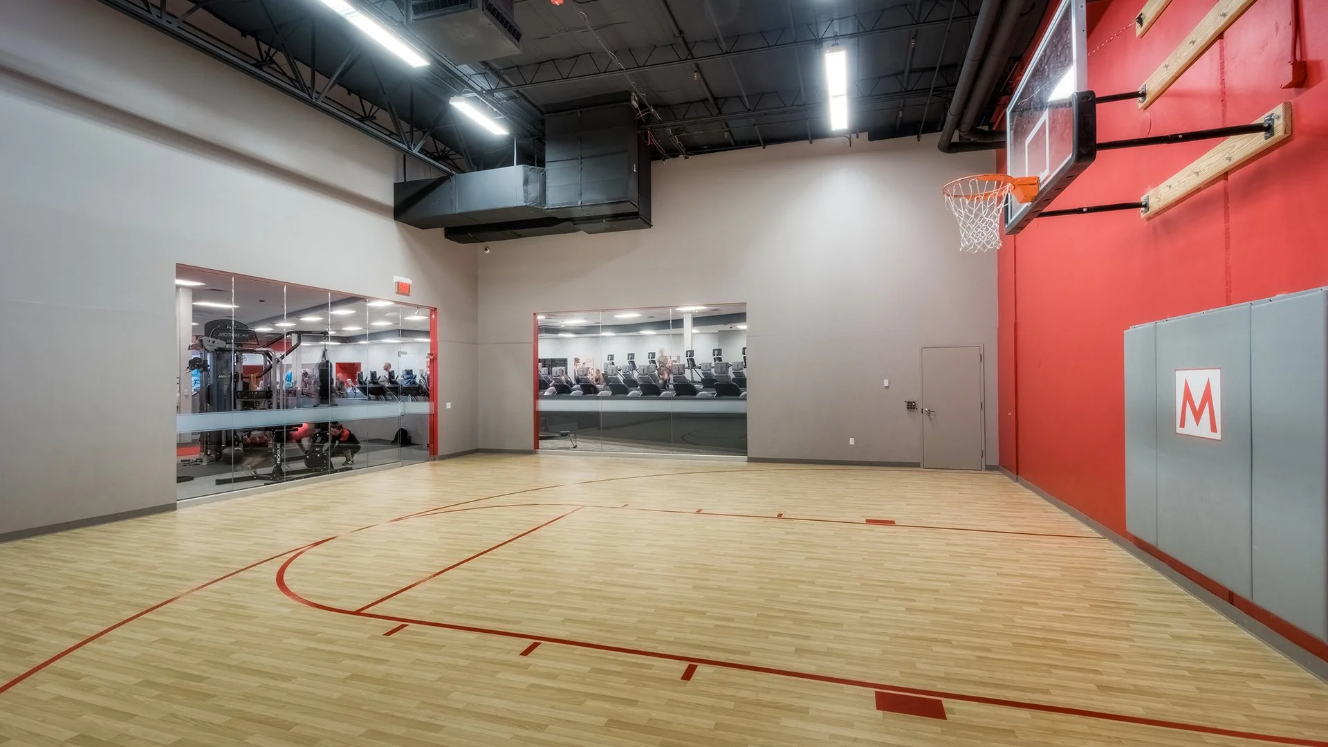 Empty indoor basketball court with wood flooring, red lines, and a hoop on the wall, adjacent to a gym with cardio equipment visible through glass windows.
