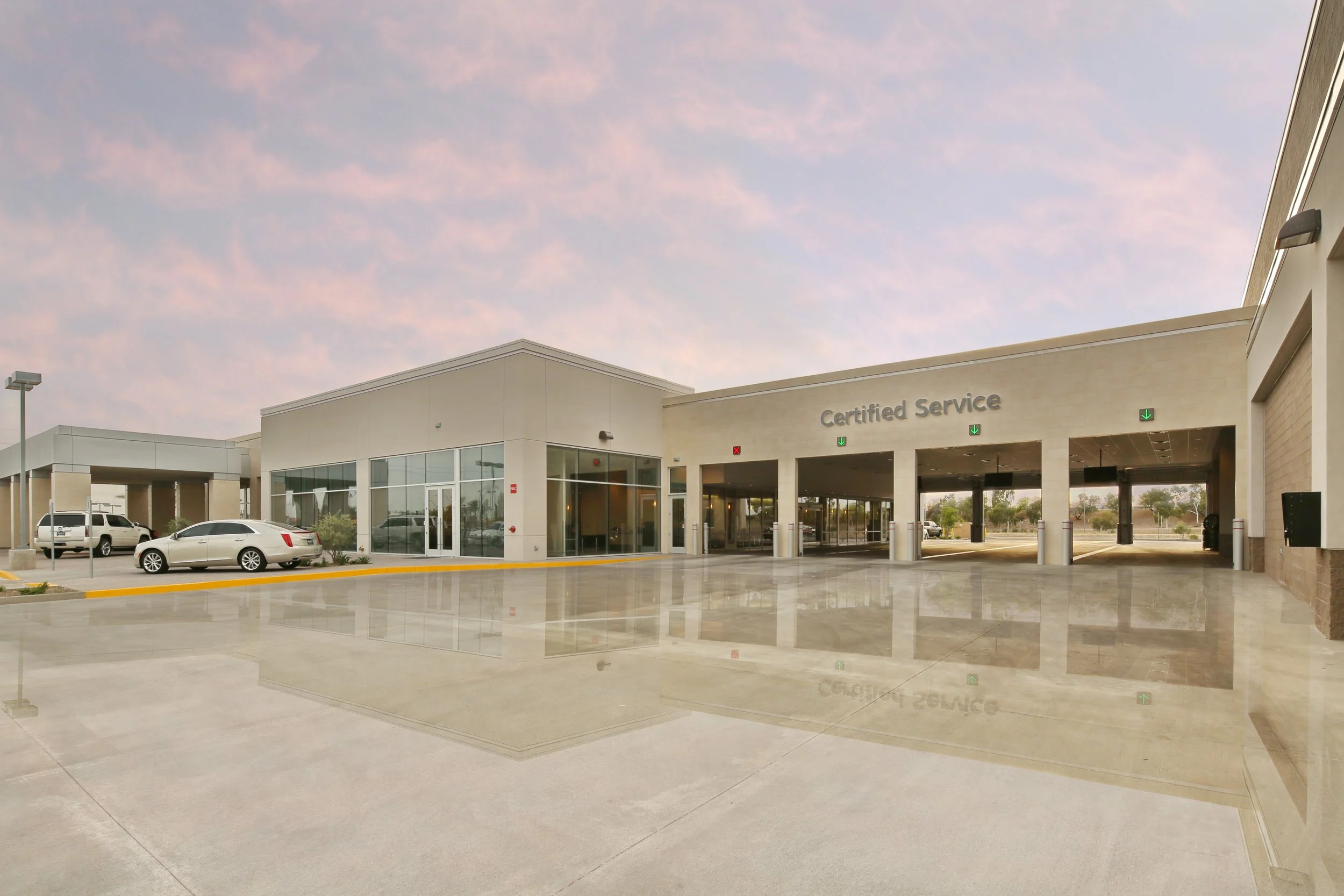 A modern car dealership with a large glass showroom and a covered certified service area. Several cars are parked outside, and the parking lot has a polished, reflective concrete surface. The sky is partly cloudy with soft pink and blue hues.