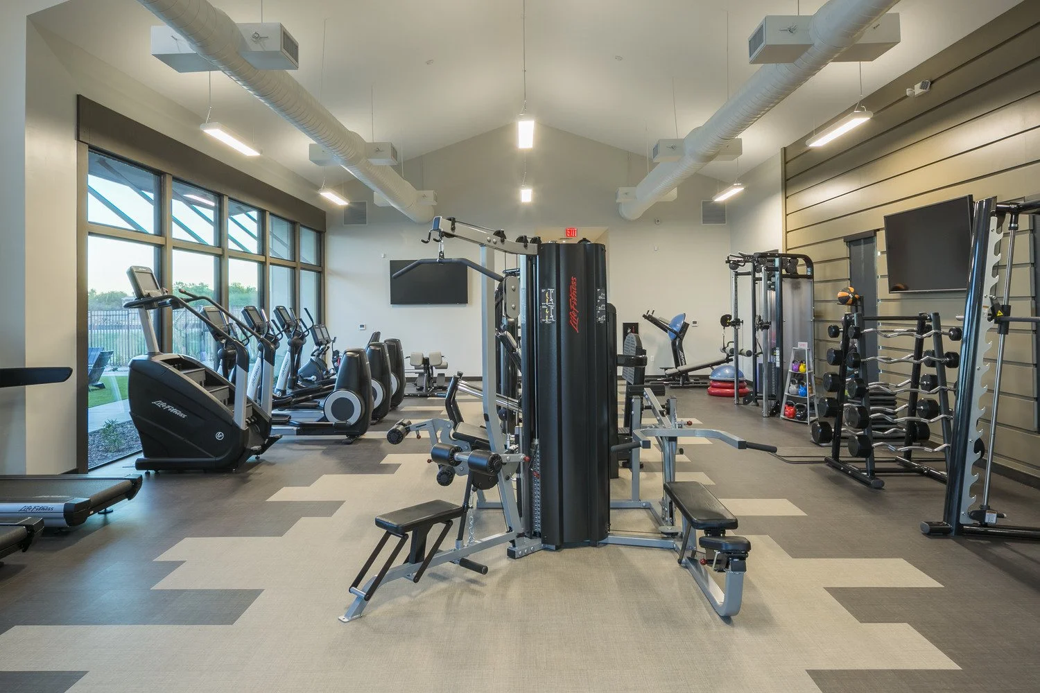 Interior of a modern gym with various exercise equipment including treadmills, ellipticals, and weight machines, with large windows and bright lighting.