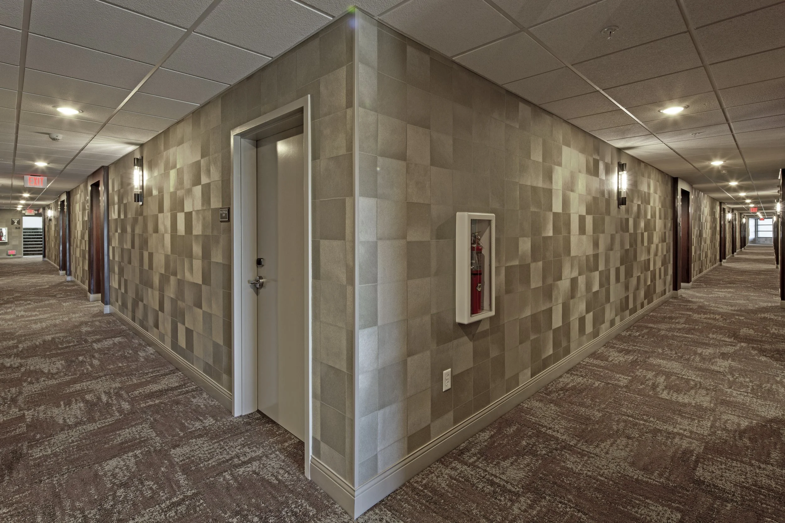 Long hotel hallway with beige tiled walls, patterned carpet, and multiple doors on both sides, ceiling lights, and a fire extinguisher inside a wall-mounted box.