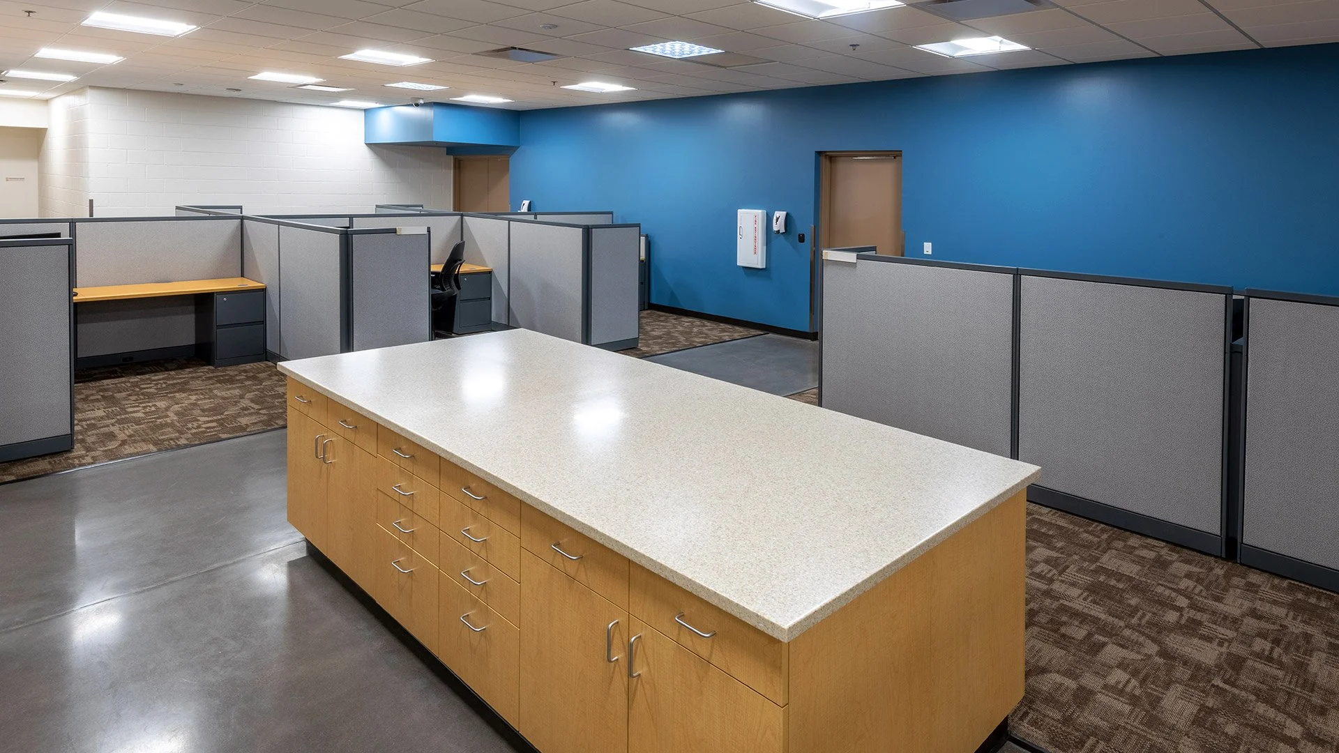 Empty office cubicles with gray partitions, a wooden table, and a blue accent wall with a door and a wall-mounted emergency box.
