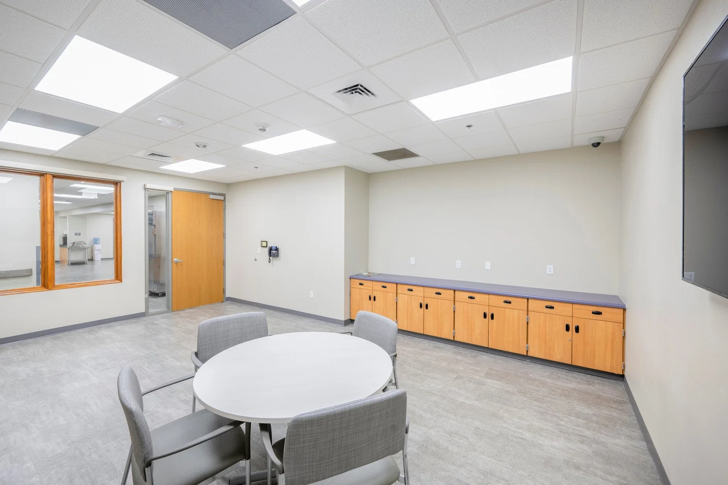 Empty conference room with round table and four chairs, wooden cabinet along wall, door, window to other room, wall-mounted TV, and ceiling lights.