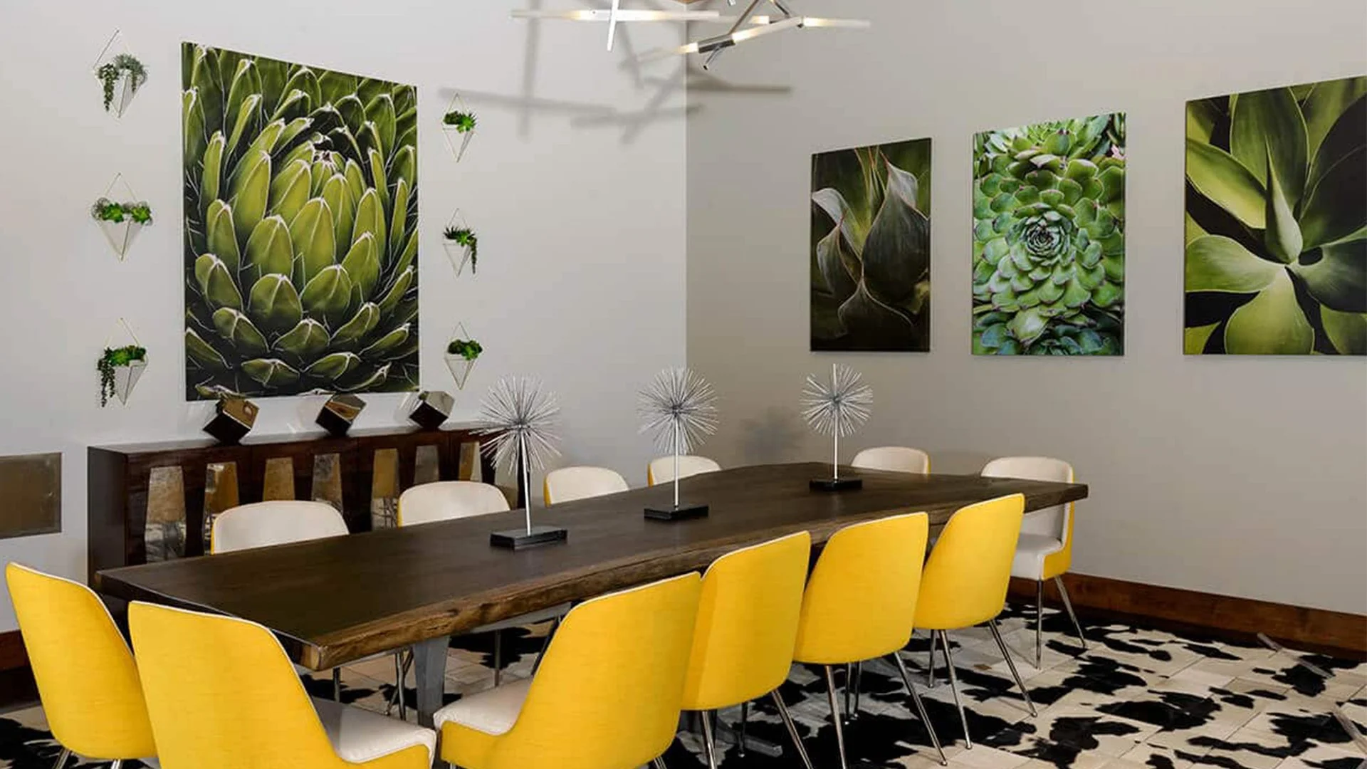 Modern dining room with a large wooden table, yellow and white chairs, decorative metal sculptures, and botanical artwork on the walls.