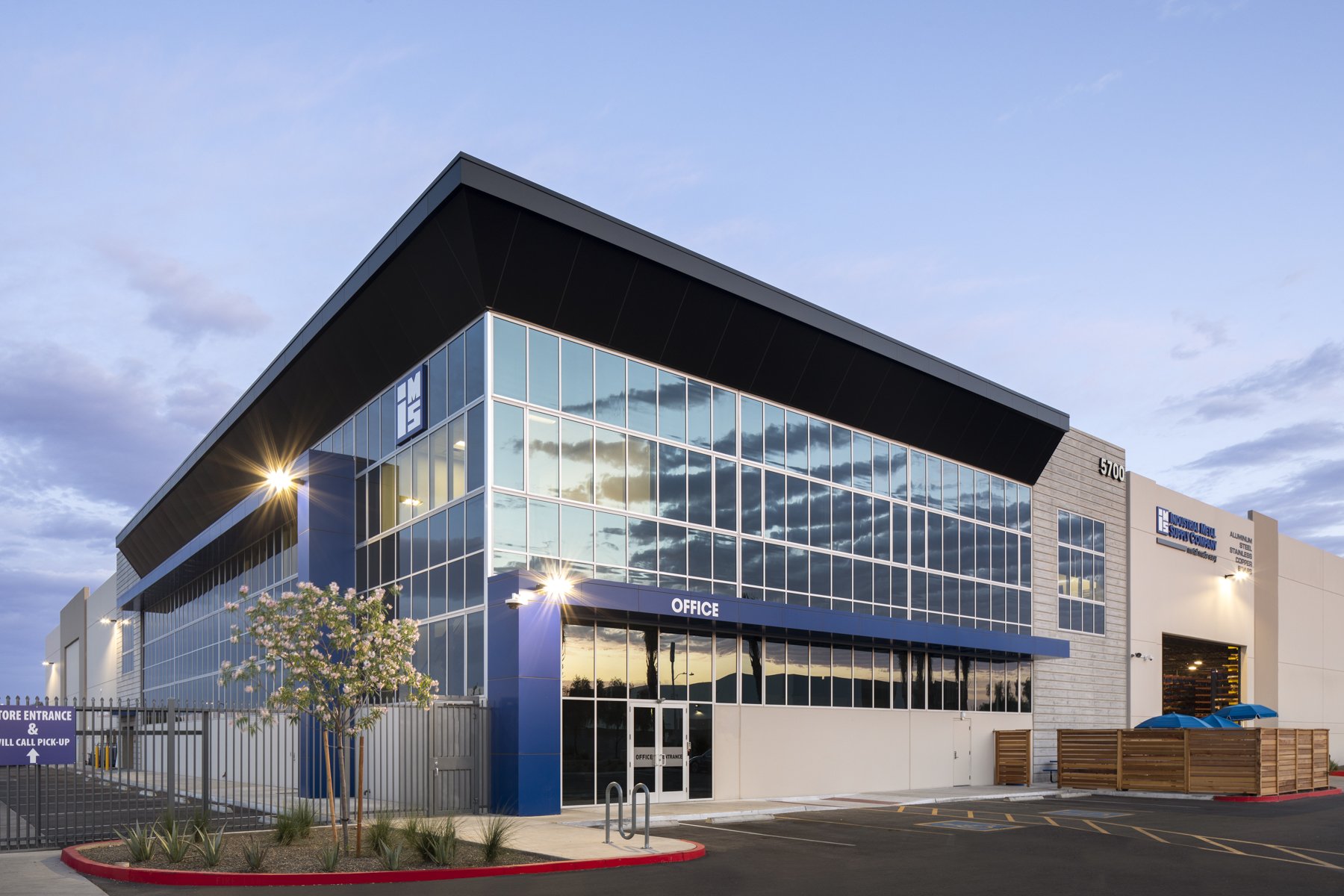 Modern office building with large glass windows, a blue sign labeled 'Office' at the entrance, a parking lot, and a small landscaped area with a flowering tree and plants.