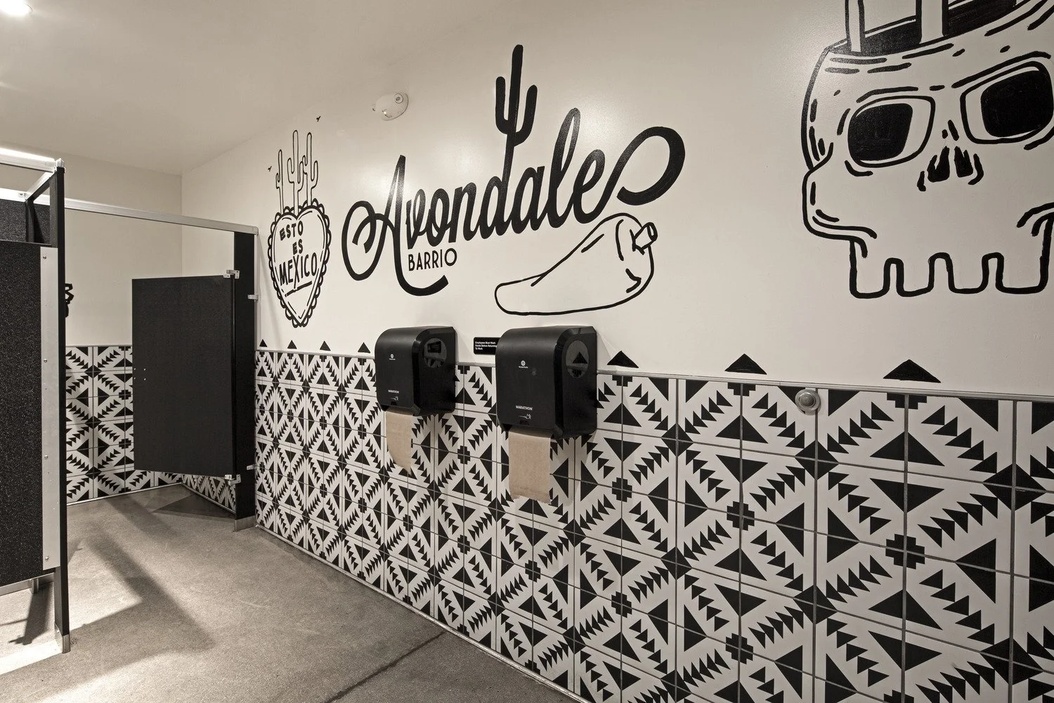 Interior wall with black and white Mexican-themed murals, including a cactus heart with the words 'Esto es Mexico,' the word 'Ayondale,' a chili pepper, and a skull wearing sunglasses, with two black paper towel dispensers and patterned tiles on the 