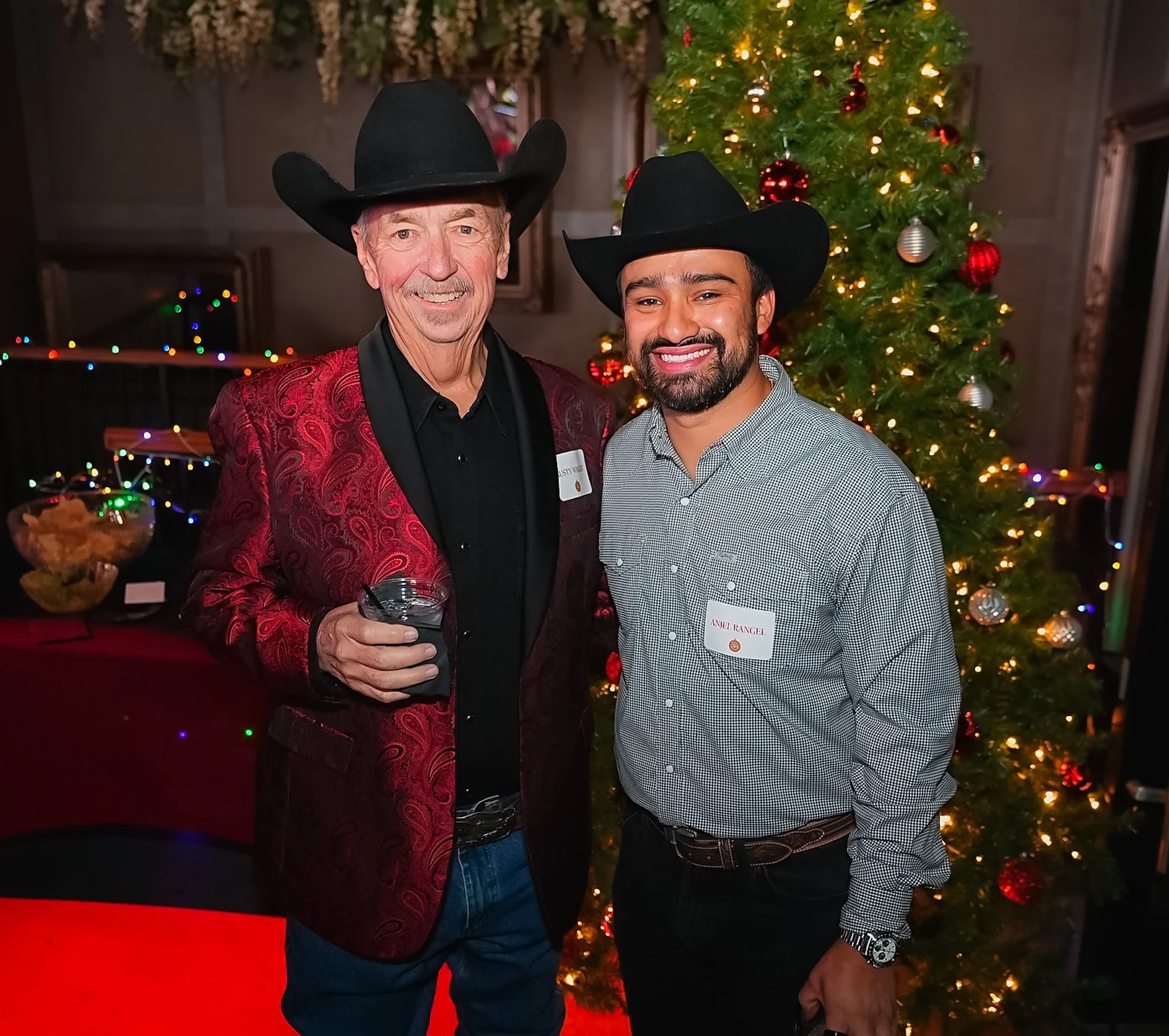 Two men wearing cowboy hats stand in front of a decorated Christmas tree, smiling at the camera. One holds a drink, and both are dressed in semi-formal attire with name tags, at a festive event.