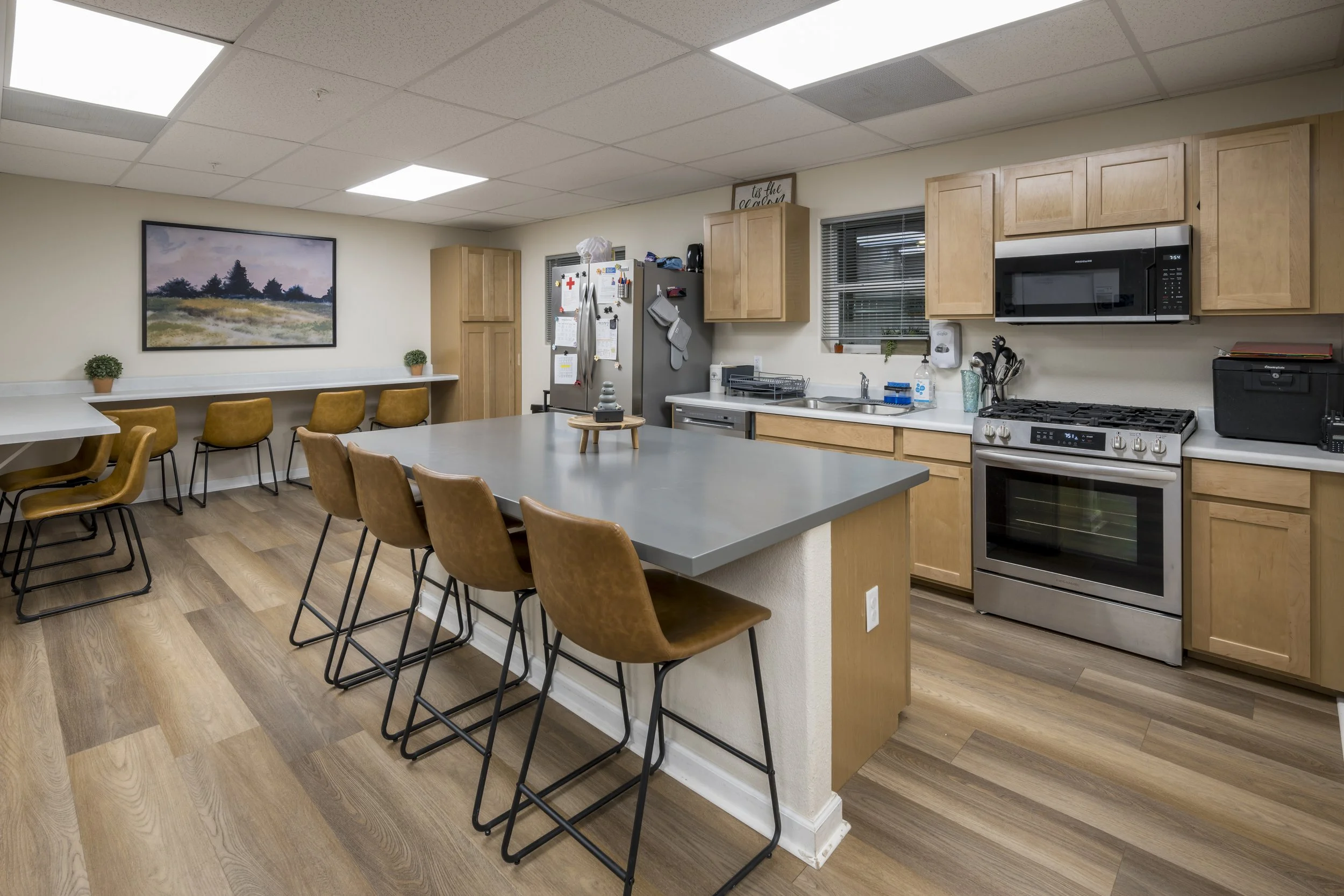 Kitchen with wooden cabinets, stainless steel stove, microwave, countertop, and bar stools, with a table and chairs in the background.