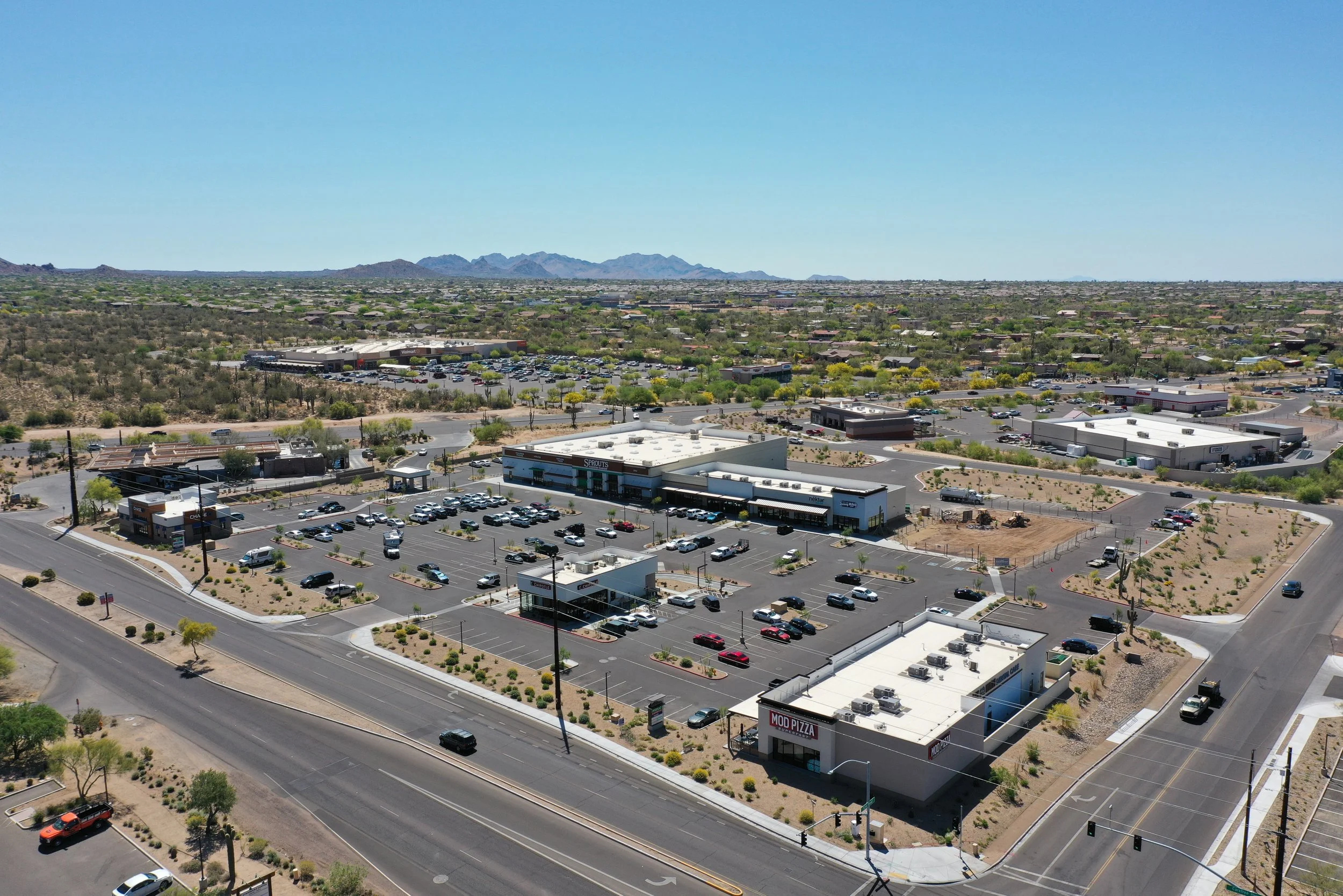 Aerial view of a shopping center parking lot and surrounding commercial buildings on a sunny day, with desert landscape and mountains in the background.
