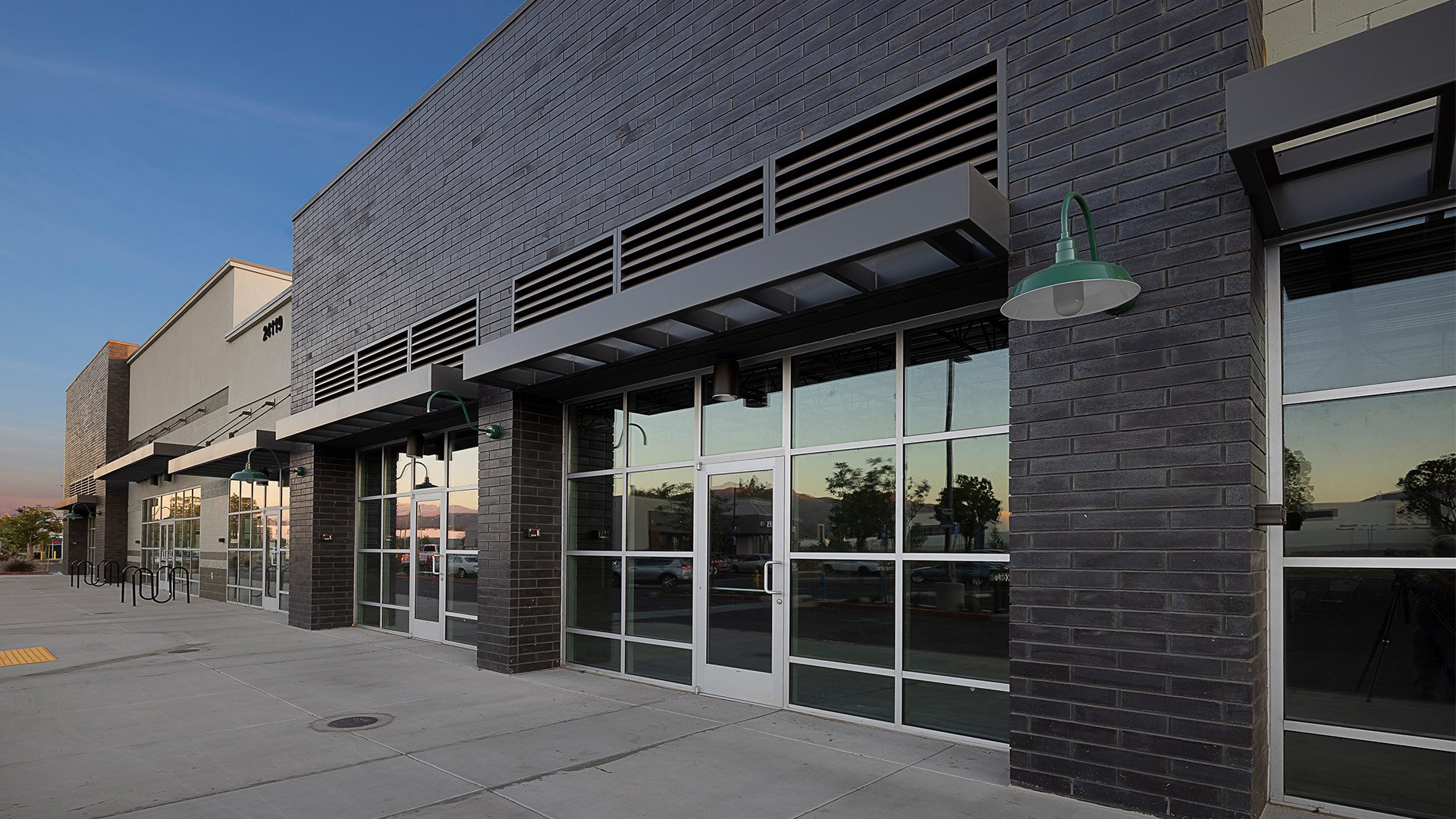 Empty storefront with large glass windows, black brick facade, green outdoor wall lights, and a sidewalk in front, during early evening.