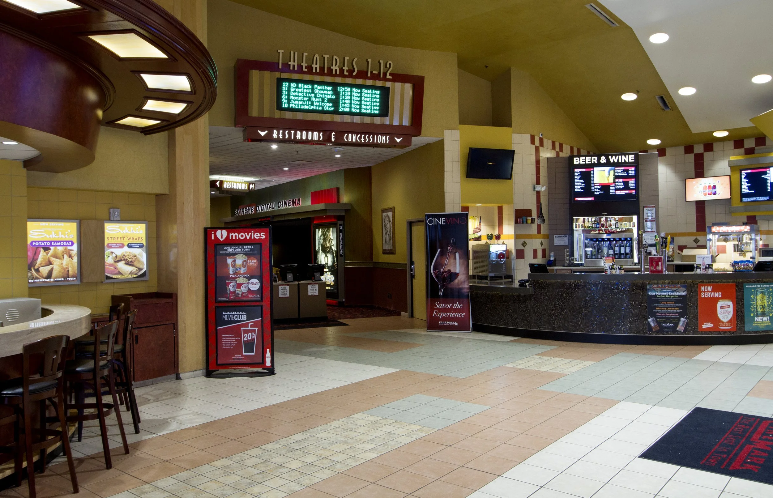 Inside a movie theater lobby with concession stand, menu boards for beer and wine, digital movie schedule, and seating areas.