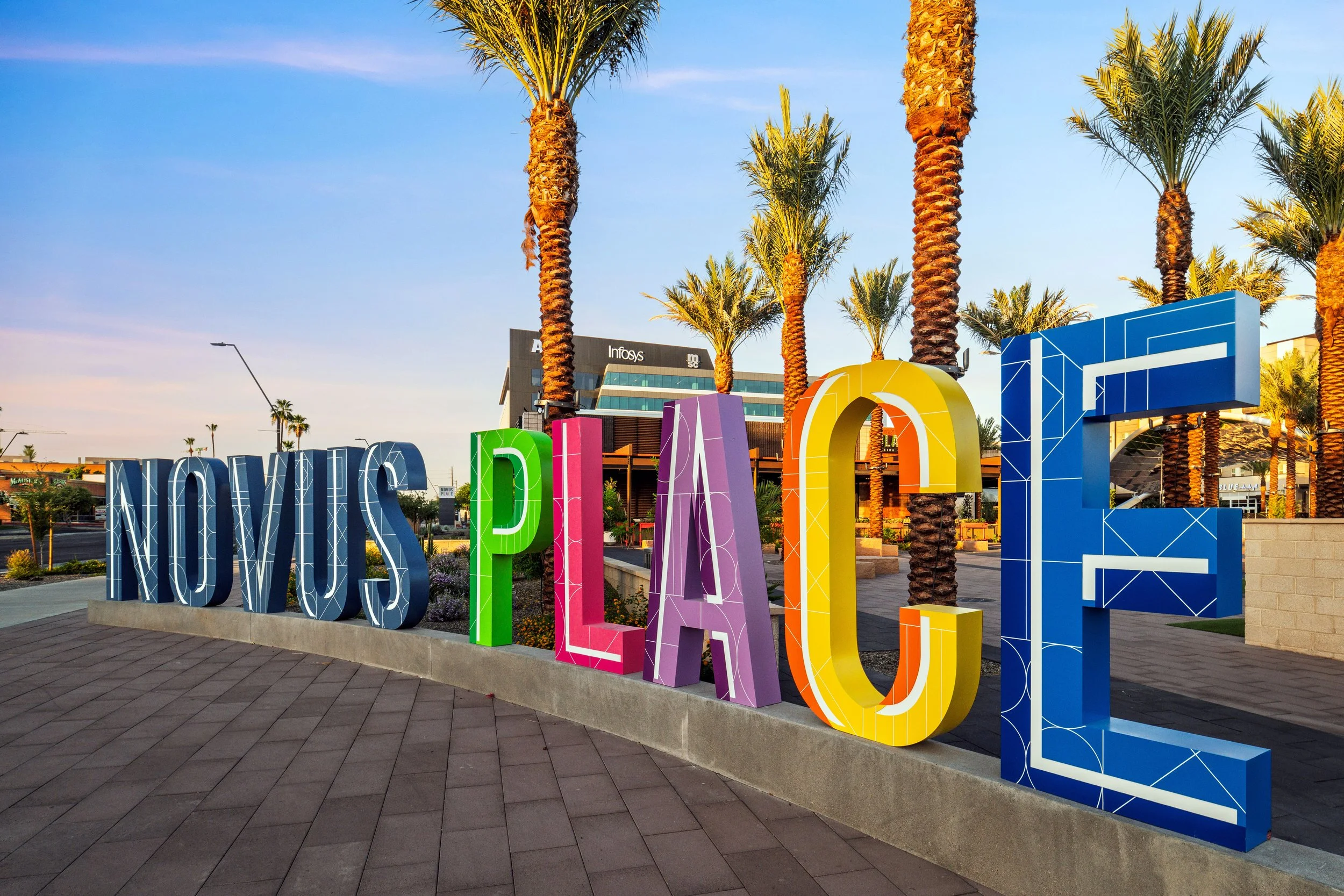 Colorful 3D sign spelling out 'Novus Place' with tall palm trees in the background, modern buildings, and a clear blue sky at sunset.