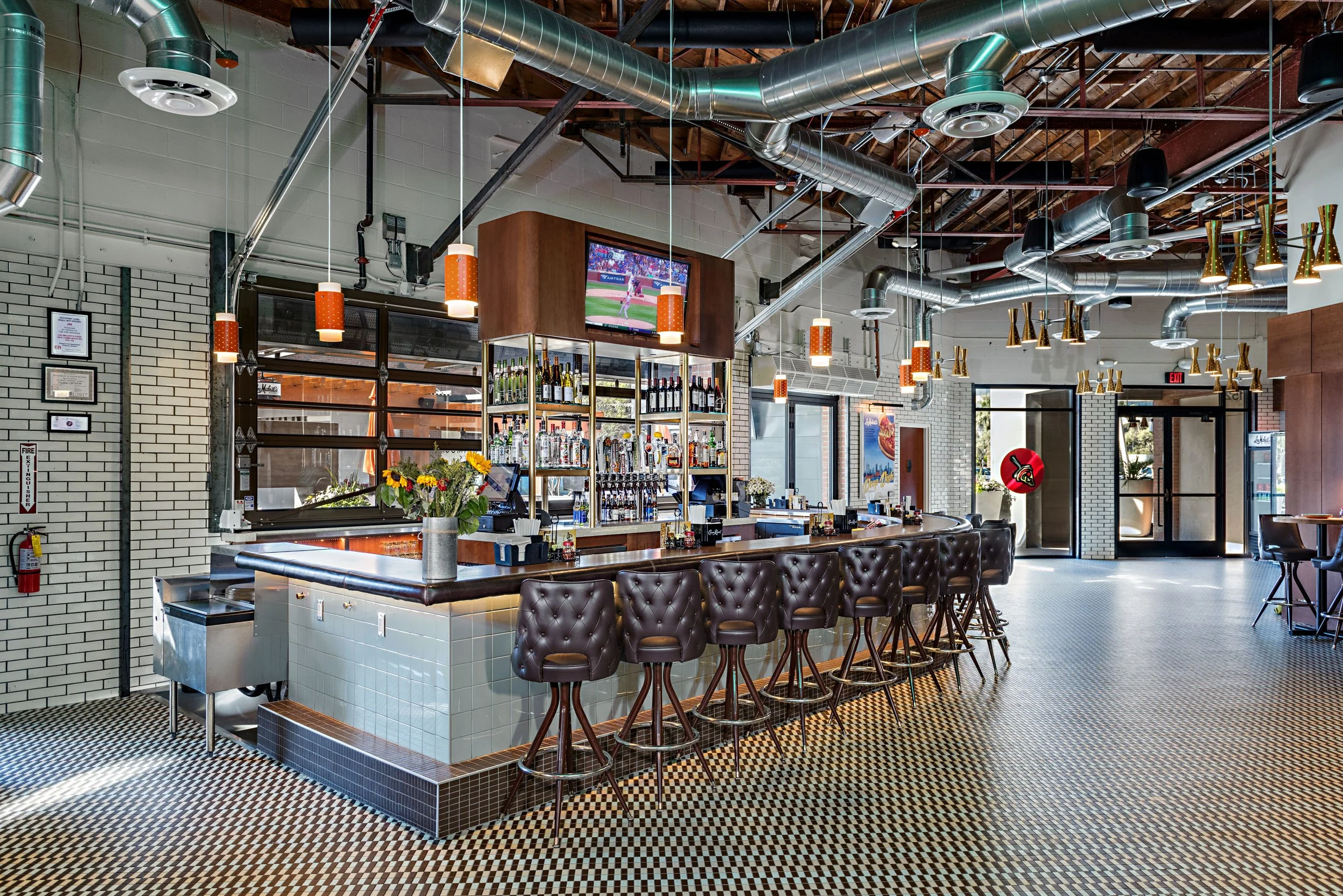 Interior of a modern bar featuring a curved bar counter with ten bar stools, industrial-style exposed ductwork, a mounted television displaying a sports game, shelves with bottles behind the bar, a flower vase, and a patterned tile floor.