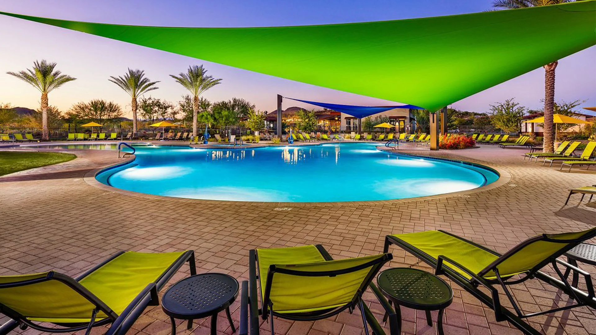 A swimming pool surrounded by lounge chairs with green cushions and large shade sails, with palm trees and buildings in the background during sunset.