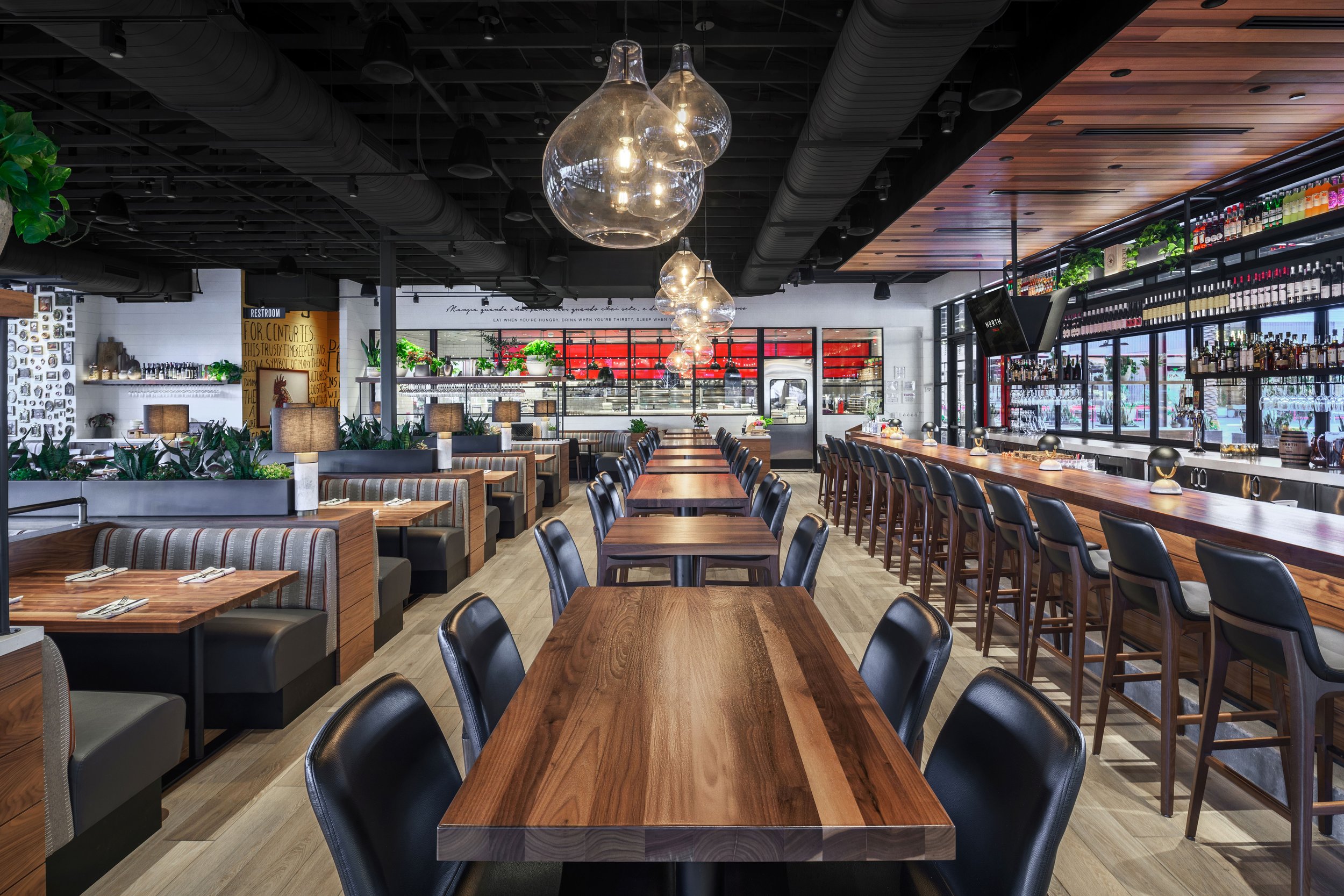 Interior of a modern restaurant with wooden tables, black chairs, hanging pendant lights, and a bar area with bottles on shelves on the right side.