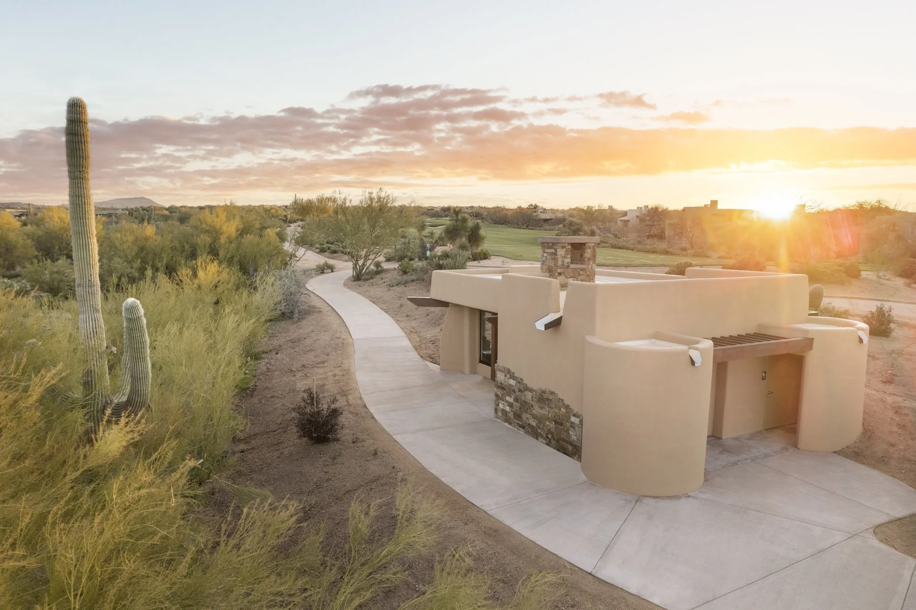 Sunset over a desert landscape with cacti, shrubs, and a modern adobe-style building with a stone accent wall and a barbecue area.